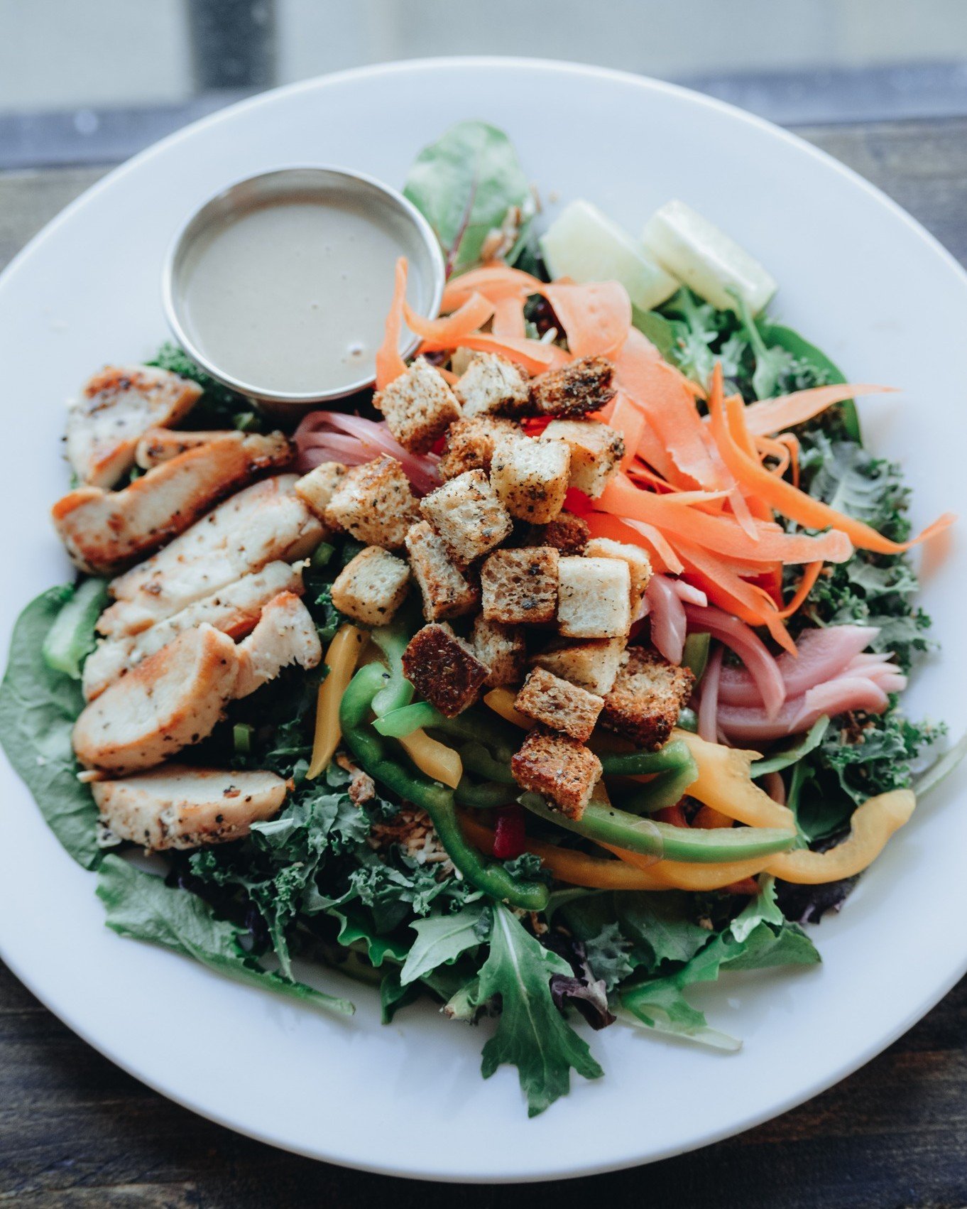 Try our delicious Thai Coconut Chicken Salad! Packed with greens, kale, chicken, bell pepper, and carrots, it also features toasted coconut for a tropical twist. Enjoy pickled onions, sourdough croutons, and a zesty lime wedge. All topped with a savo
