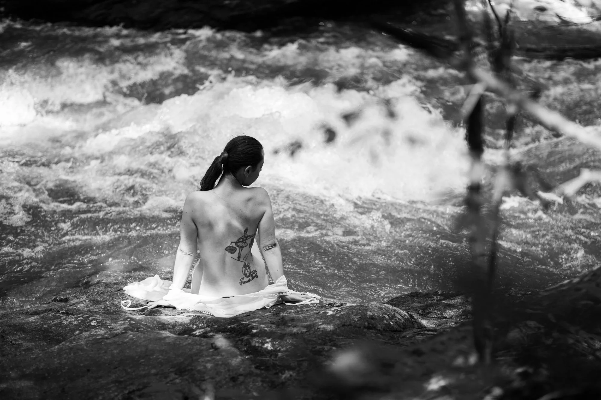 black and white boudoir portrait of a woman's back as she sits on a boulder next to a rushing river near Charlottesville, Virginia.
