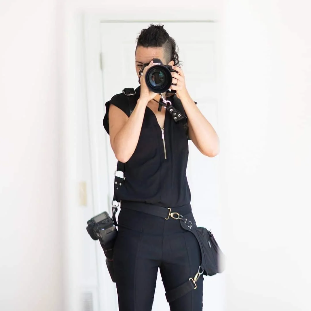 Jen Fox in a mirror holding a camera up to her face while wearing all black. A camera hangs off her side and a hip bag is belted around her waist and thigh.