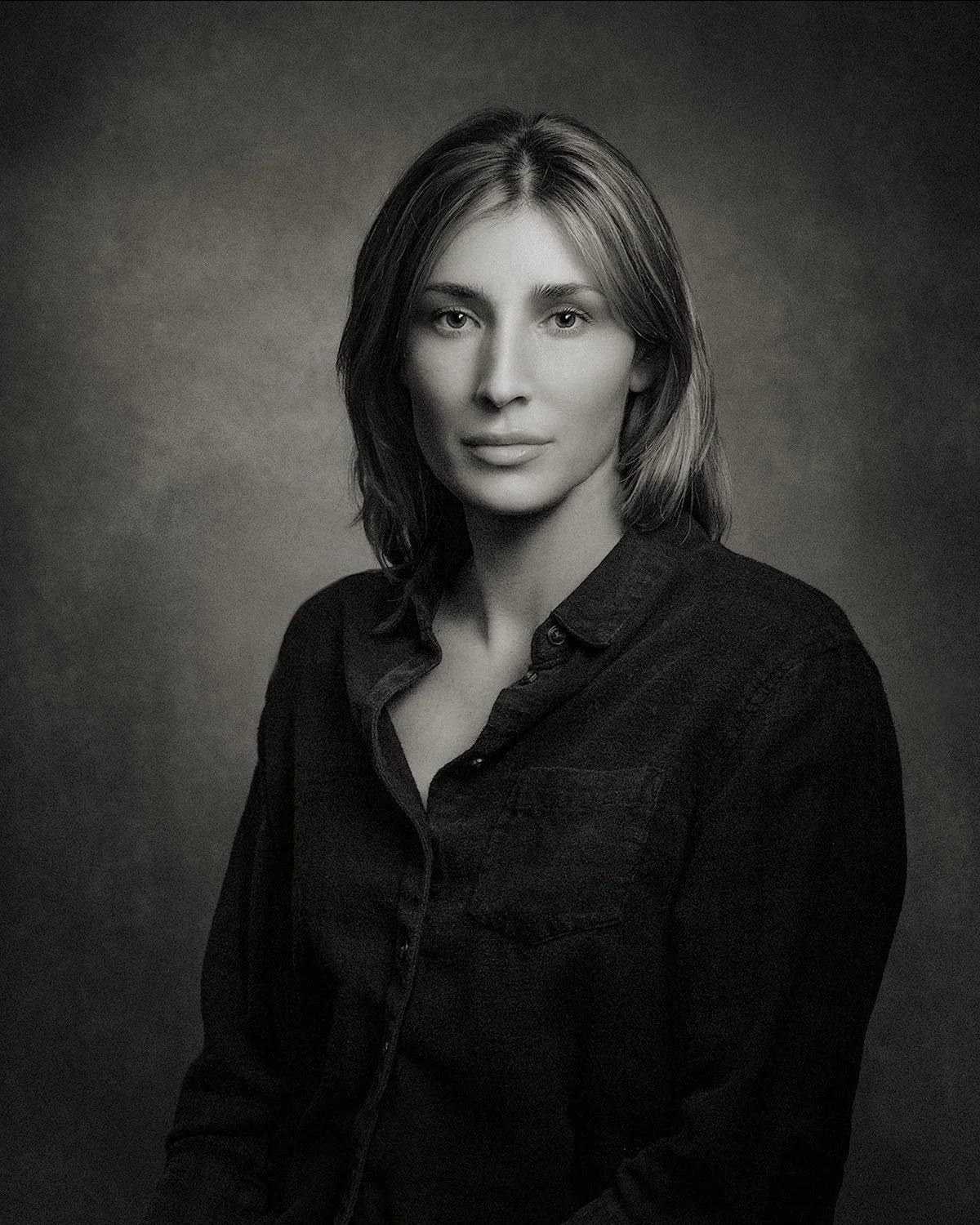 Black-and-white platinum portrait of a woman seated against a softly textured gray backdrop, photographed with quiet, natural expression. Portrait Alchemy.