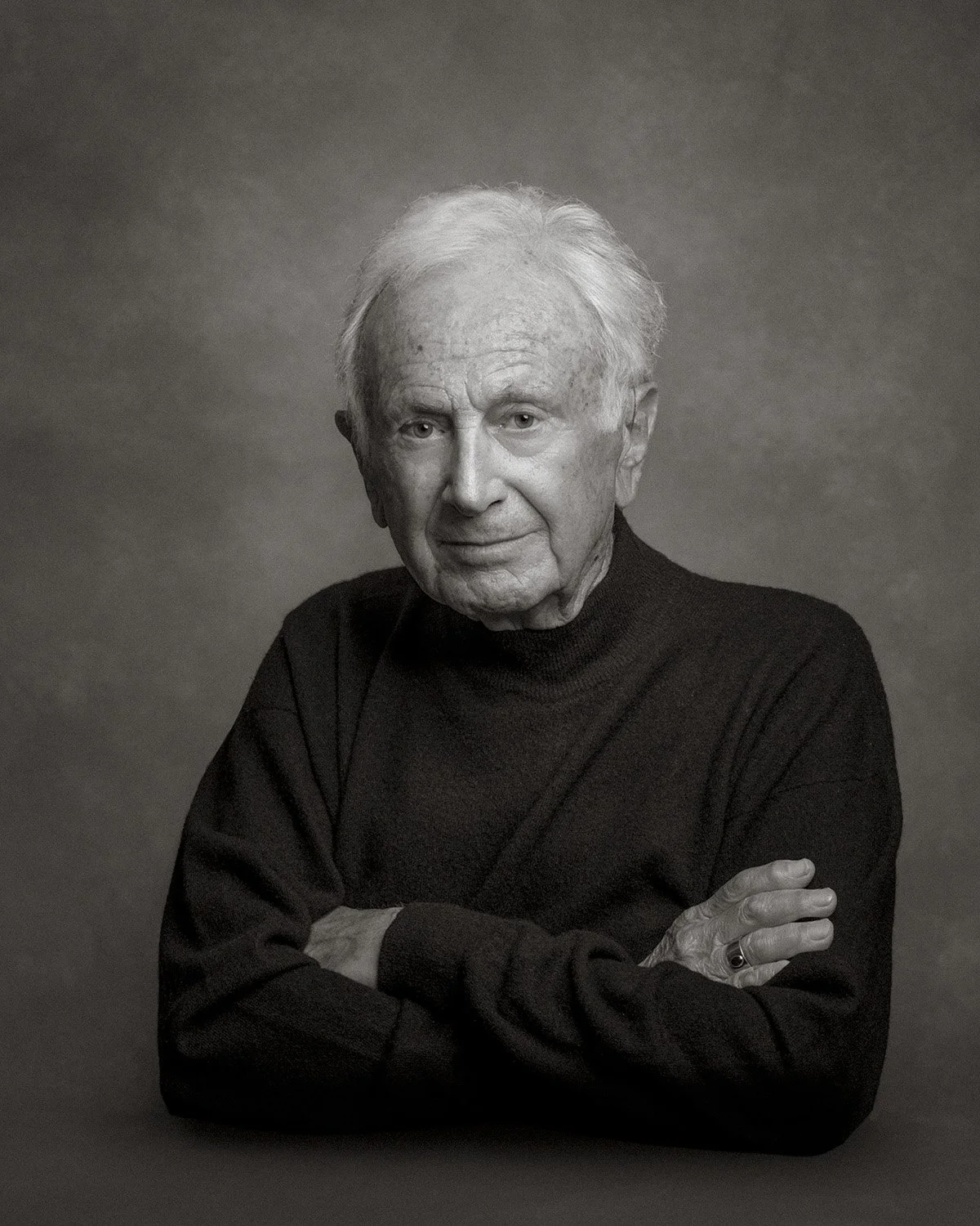 Black-and-white platinum portrait of an older man seated with arms folded, photographed against a softly textured gray backdrop. Portrait Alchemy