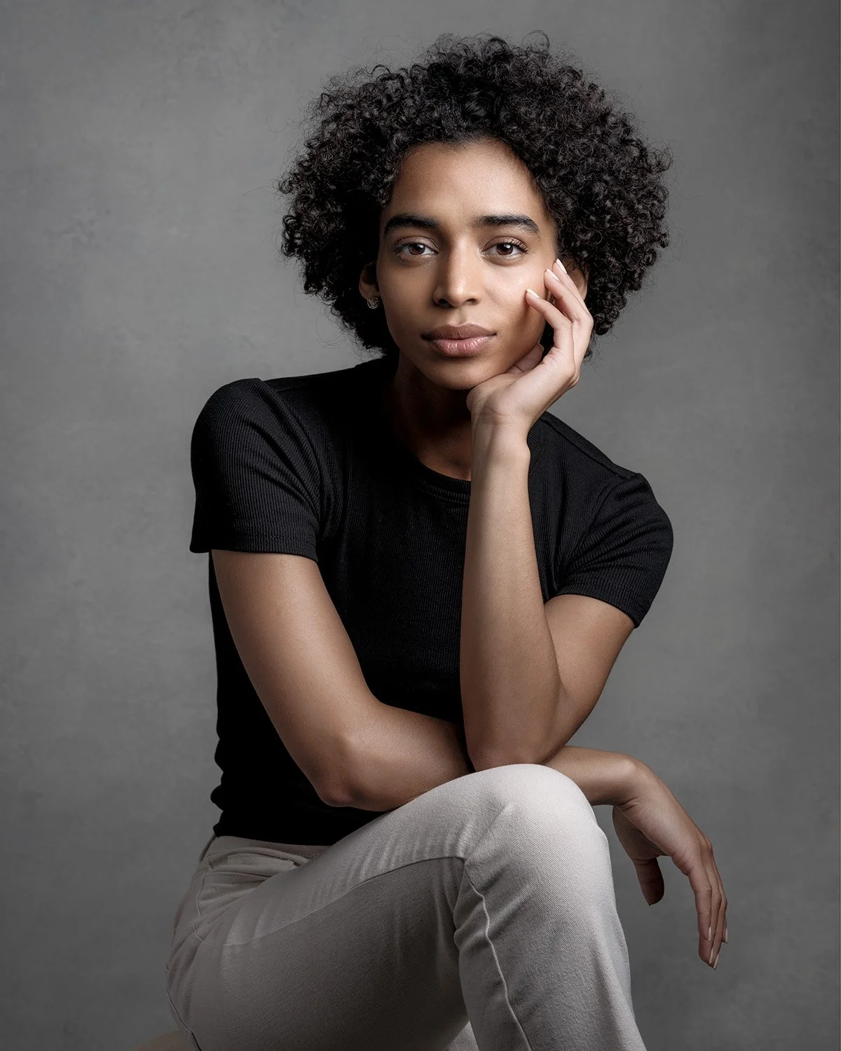 Traditional studio portrait of a young woman with natural curls, seated thoughtfully against a soft gray backdrop. Portrait Alchemy.