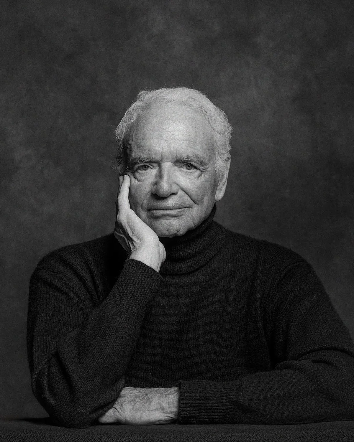 Black-and-white studio portrait of an older man resting his face on his hand, photographed against a soft gray backdrop. Portrait Alchemy.