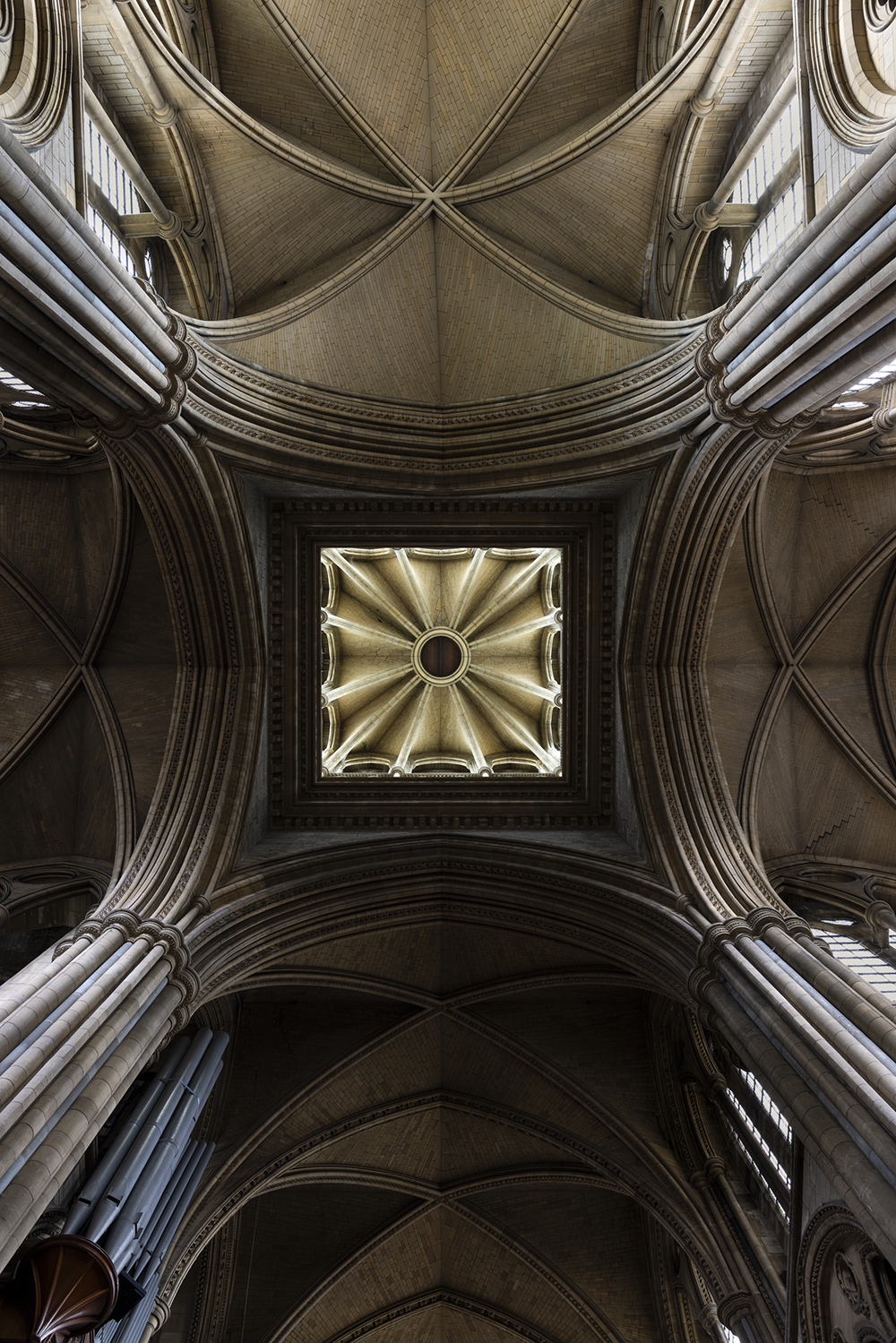 Interior view of a Gothic cathedral ceiling with intricate stone ribbed vaults and a central square opening revealing a domed ceiling with radiating ribs.