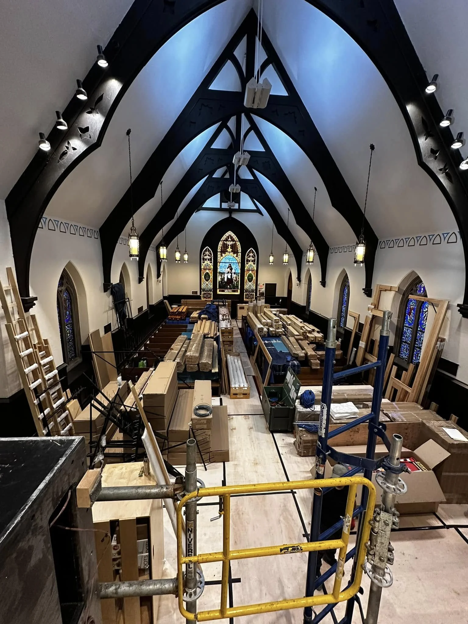 Interior view of a church sanctuary filled with construction materials, lumber, and scaffolding during a renovation project, looking toward one of our historic stained-glass windows.