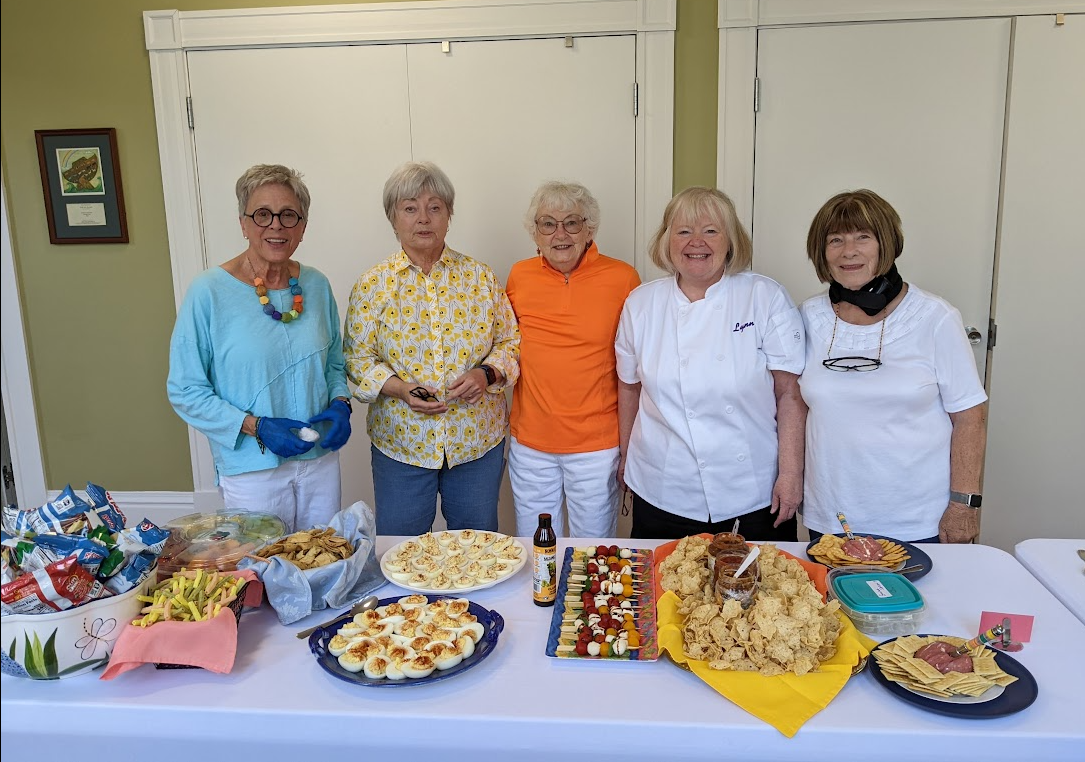 A group of 5 women stand together in front of a table full of food.