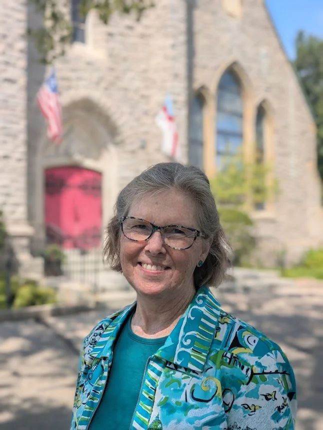Portrait of Deacon Anne Reed outdoors in front of the historic stone church with its red door in the background