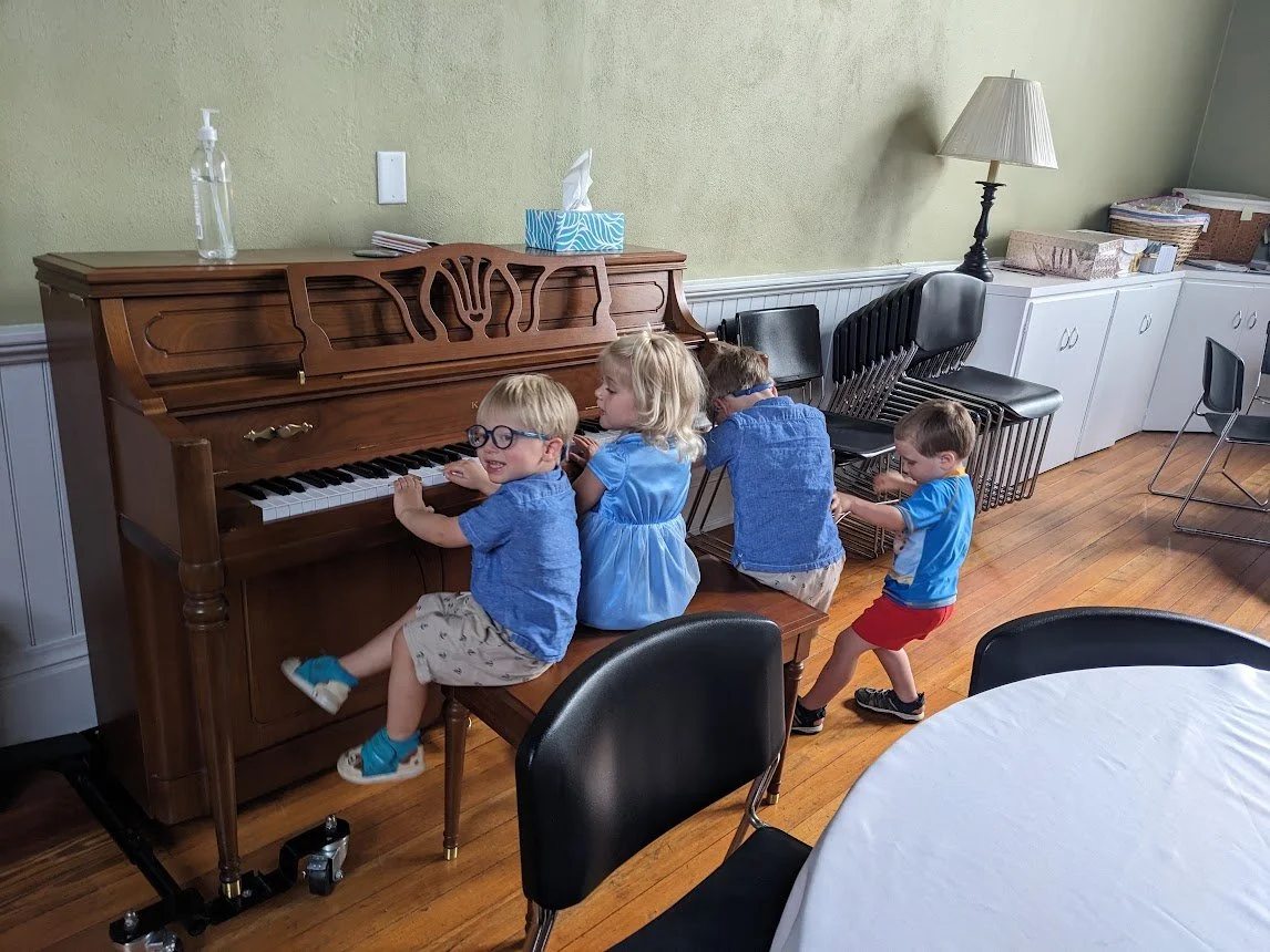 Four young children joyfully gathered along a wooden piano bench playing music together during church fellowship.