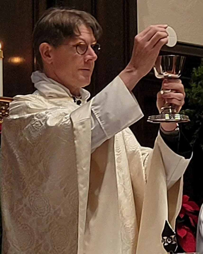 Clergy member lifting the communion wafer and silver chalice during a worship liturgy.