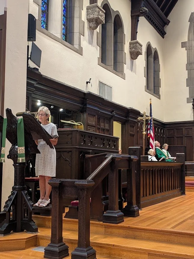 A woman stands at a dark wood eagle lectern reading during an Episcopal worship service, with choir seating, clergy, and Gothic architectural details in the background.