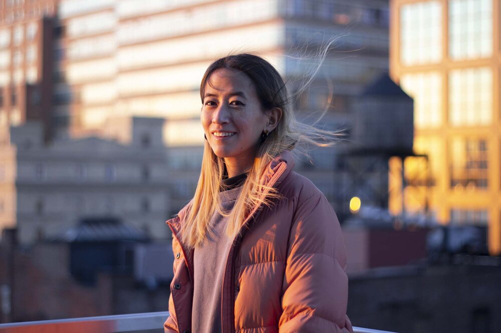 Yo-Yo Lin is east-Asian with black hair dyed bleach blonde at the ends, wearing a pink puffer jacket and smiling in an orange pink light. Her long hair is flowing in the wind on a rooftop in New York City at sunset.