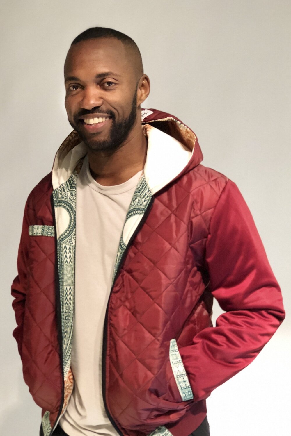녹색, 주황색, 흰색 패턴 안감이 있는 빨간색 새틴 후드 재킷을 입은 흑인 남성의 상체 샷으로, 그의 손은 주머니에 있으며, 그는 카메라를 편안하게보면서 미소짓고 있습니다.
