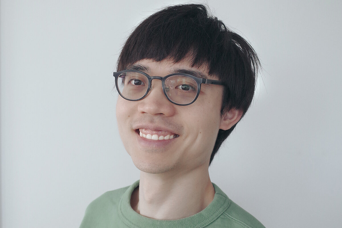 An man smiles at a three quarter angle to the camera againsta  white wall. He wears black rimmed glasses and a green crew neck sweater.jpg