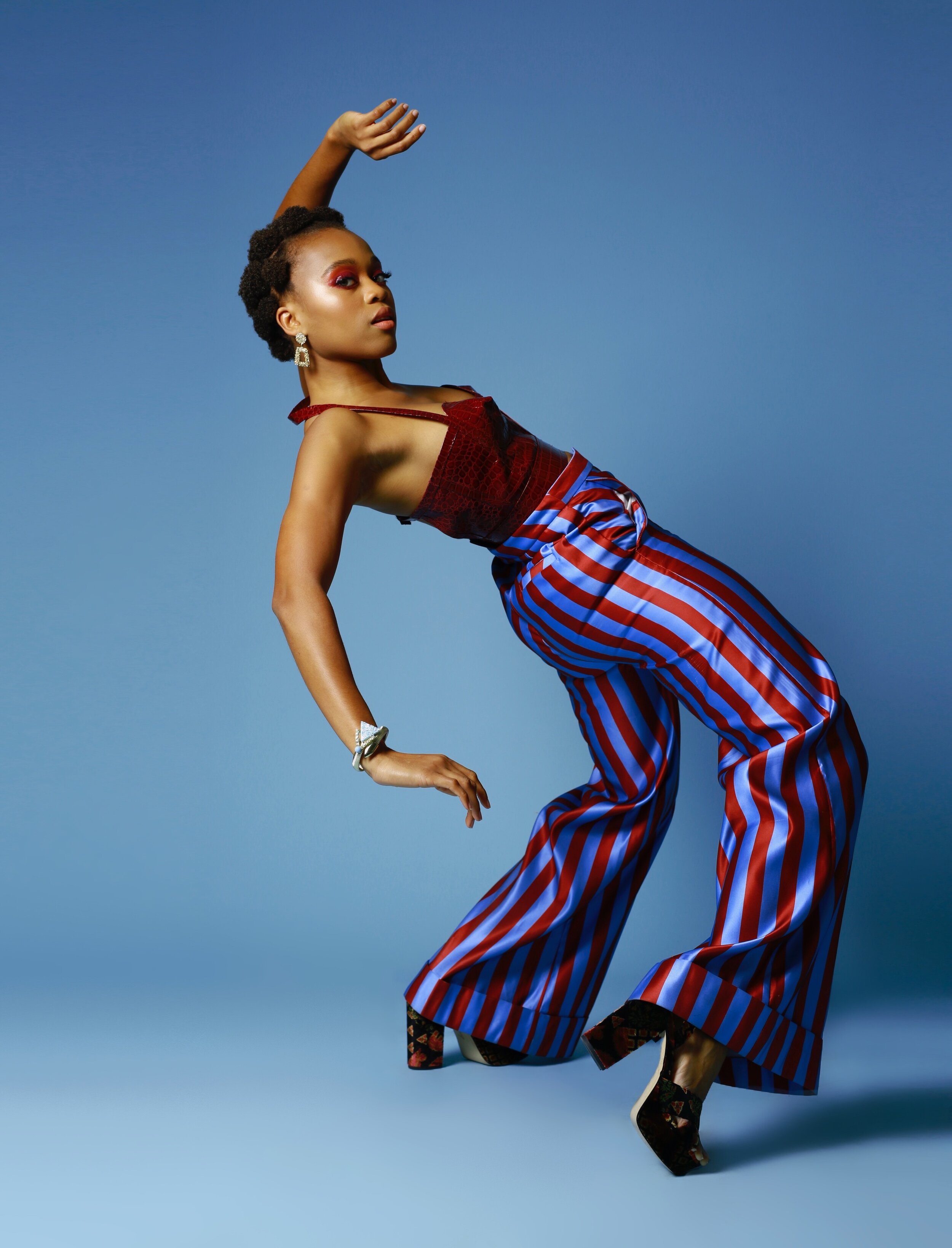 A Black woman is caputred mid-back bend in a graceful, powerful position with one hand up and one hand down. She wears a red tank top and shiny blue purple red striped bell bottom pants with chunky high heels.jpg