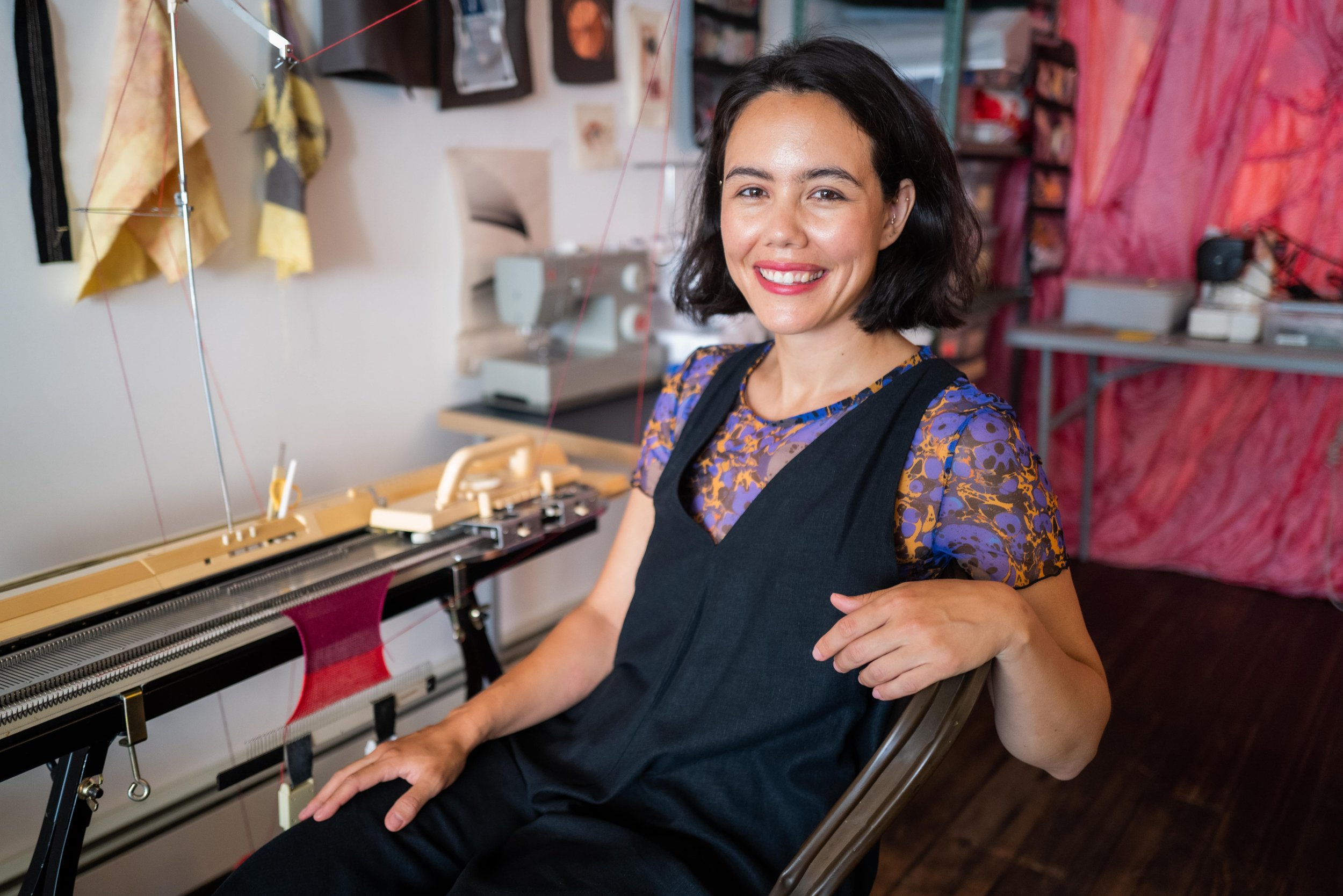 Headshot of Nicole Yi Messier wearing a black jumpsuit and purple top, sitting in front of her knitting machine at her studio.