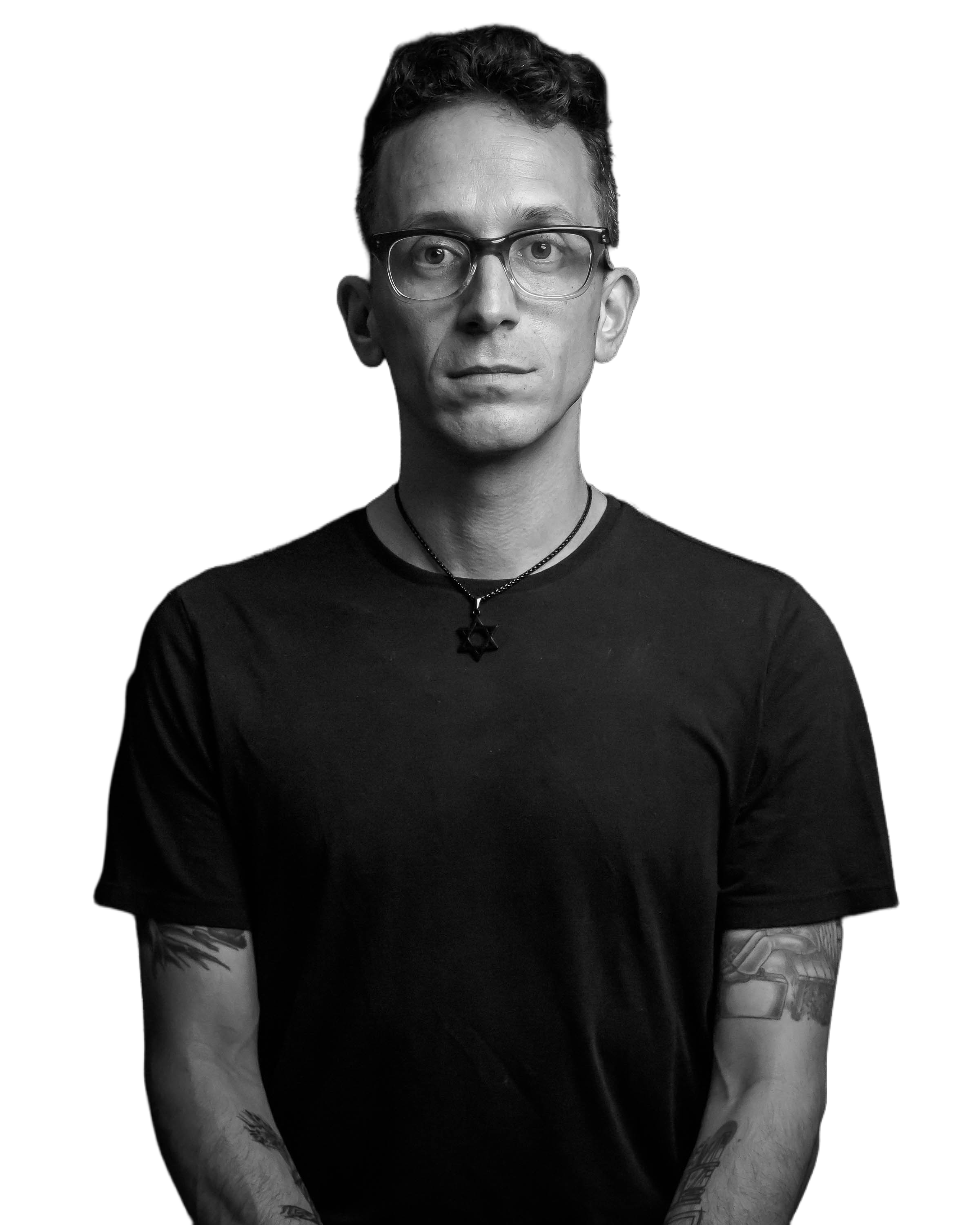 Black and white photo of a young man with glasses, wearing a black T-shirt, with tattoos on his arms and a star-shaped pendant necklace, against a black background.