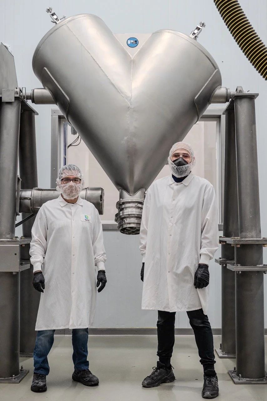 Two manufacturing workers wearing safety and sanitation gear by the V-blender