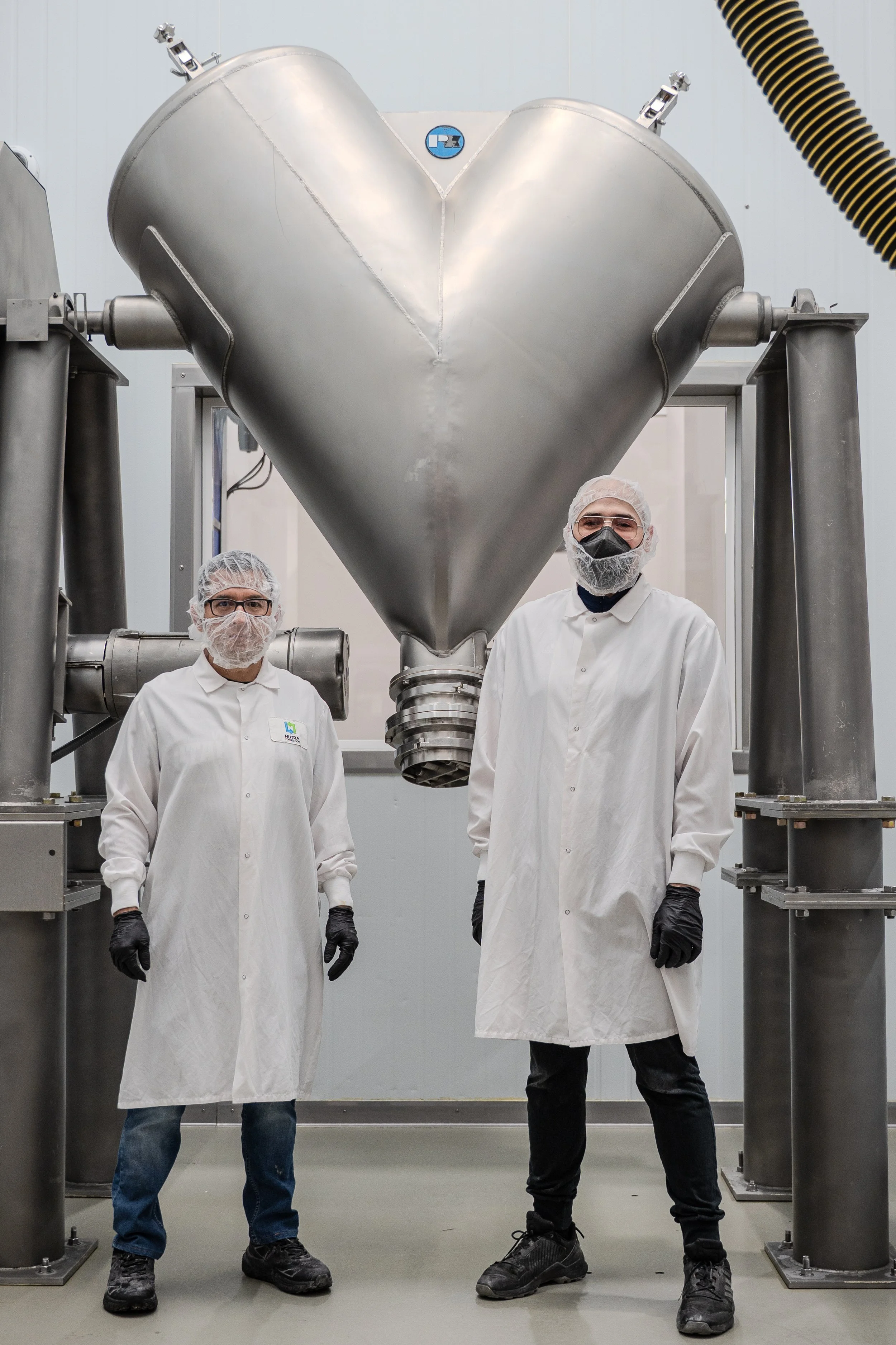 Two manufacturing workers wearing safety and sanitation gear by the V-blender