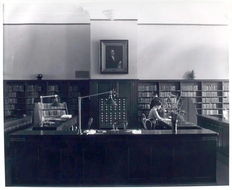 black and white photo from 1875 of a reference librarian seated at a large desk. behind the desk are bookshelves and a card catalogue with small drawers, as well as a painting hanging high on the wall of a man