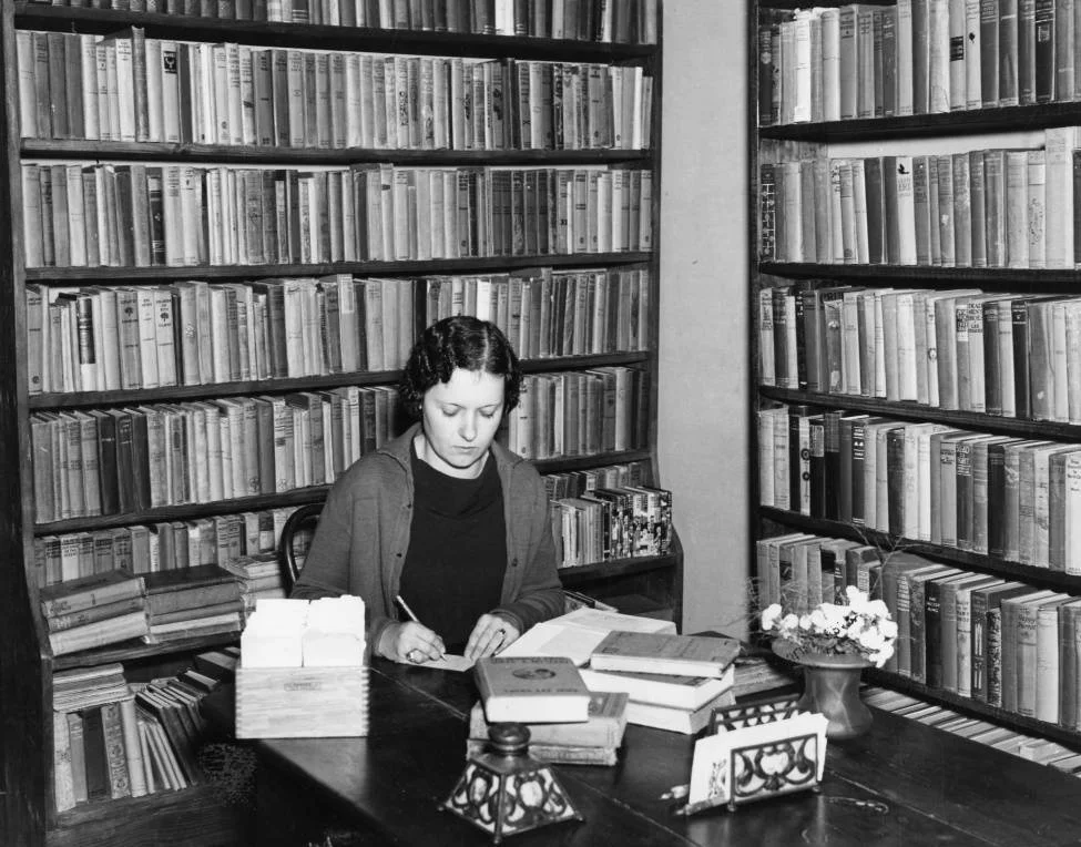 woman sitting in a library surrounded by bookshelves and books on a table in front of her, looking down, writing on a piece of paper