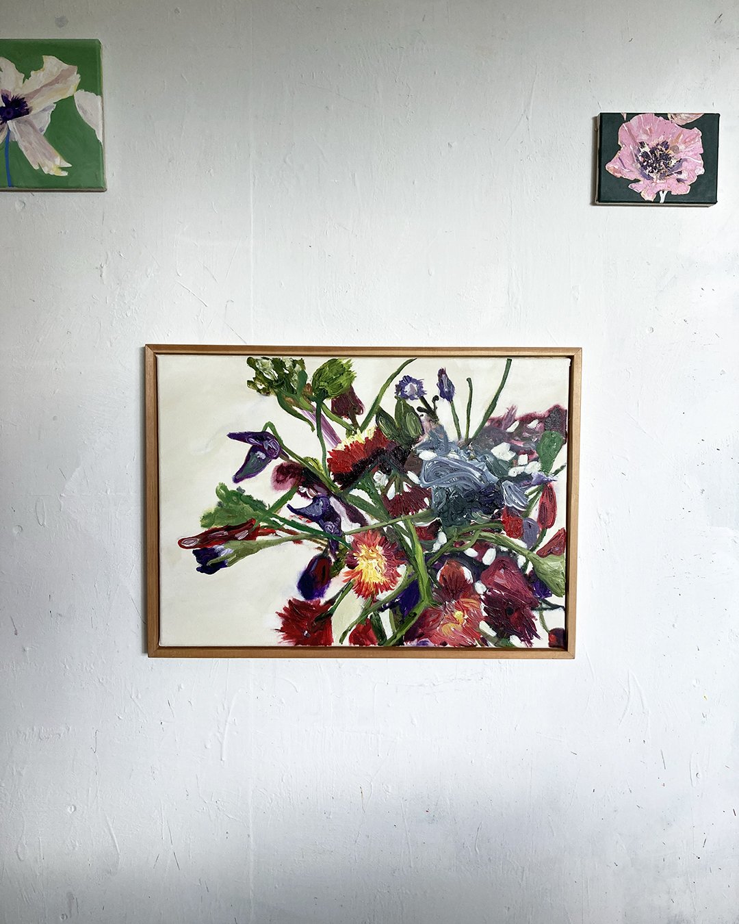A framed, colorful painting of a flower bouquet on a white wall, with two smaller floral paintings above it.