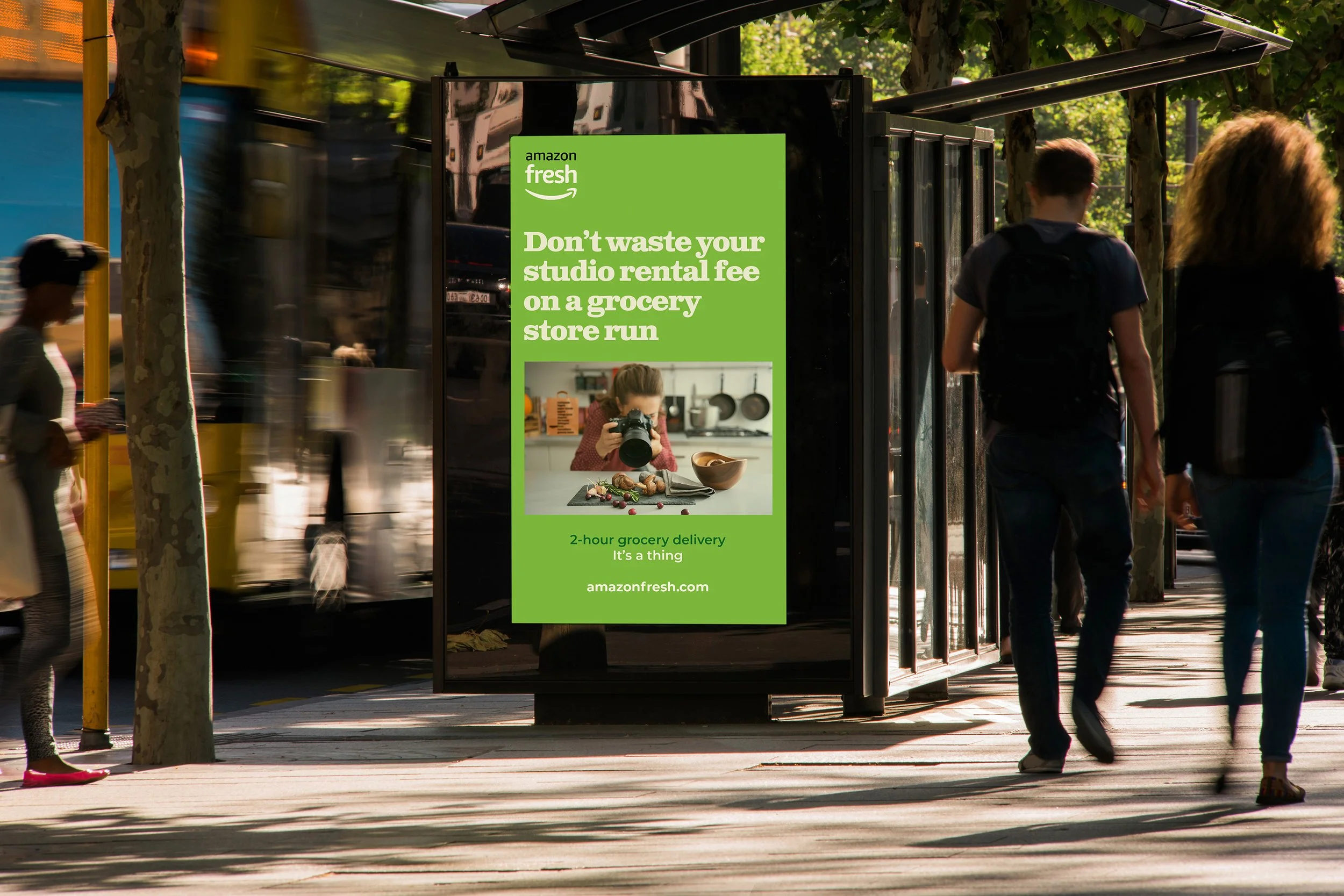 An outdoor bus shelter display showing a green Amazon Fresh advertisement targeting photographers, set in a busy street.