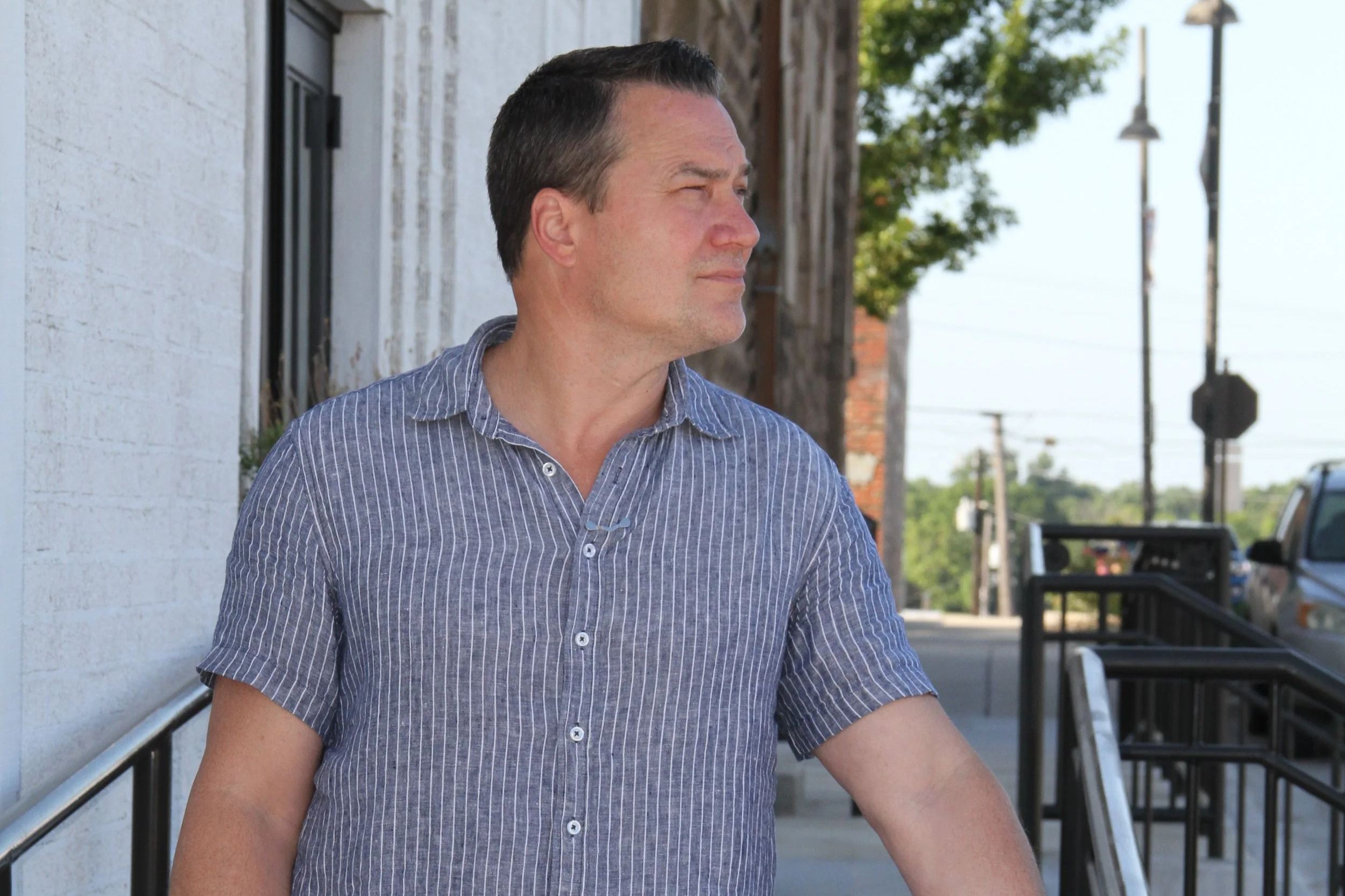 A man with dark hair wearing a blue and white striped shirt, standing outdoors in front of a white wall and some trees, with a few streetlights and cars in the background.