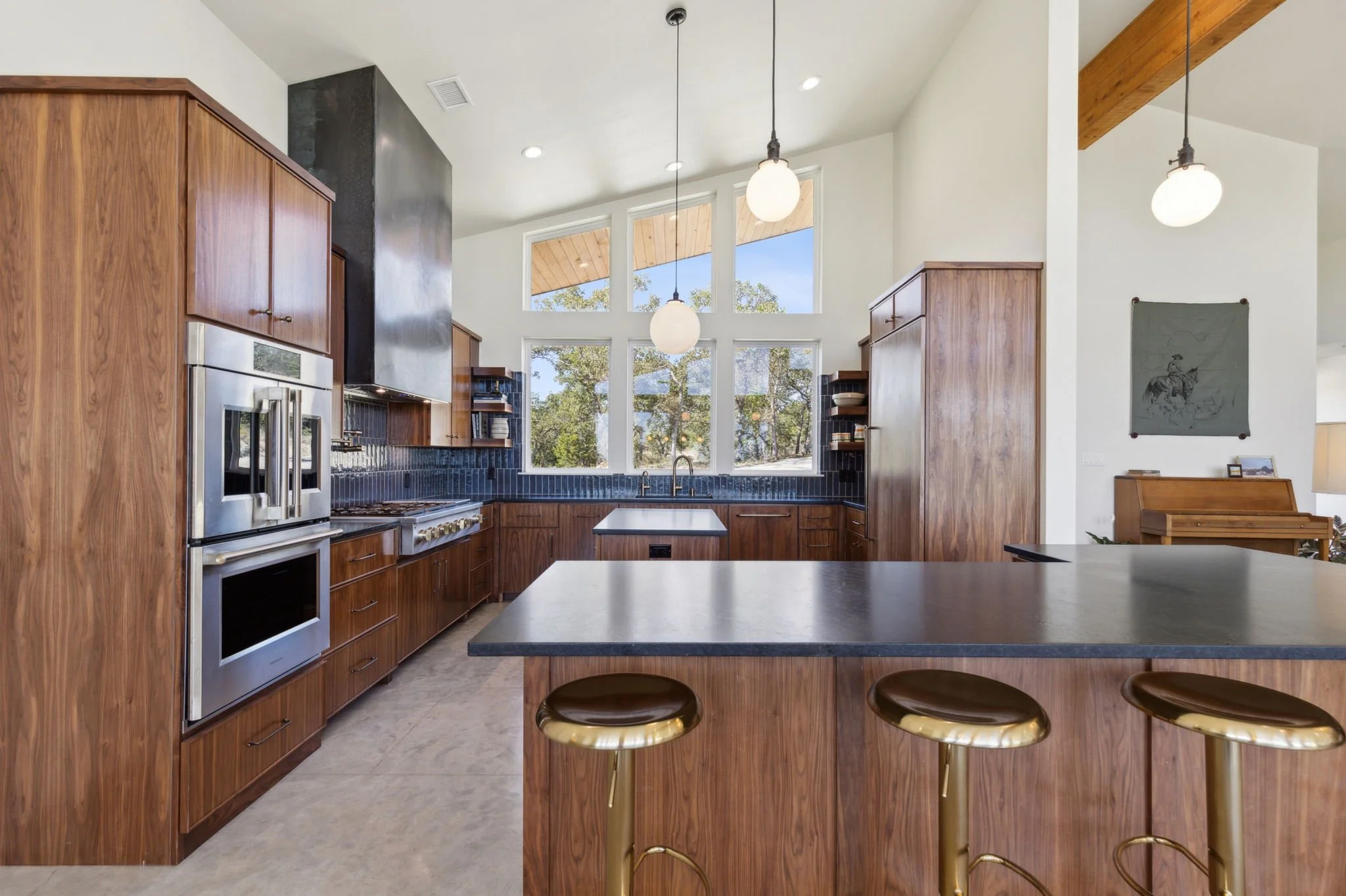 Modern kitchen with wooden cabinets, black countertops, and large windows with a view of trees outside. There are three hanging lights and a kitchen island with three gold bar stools.