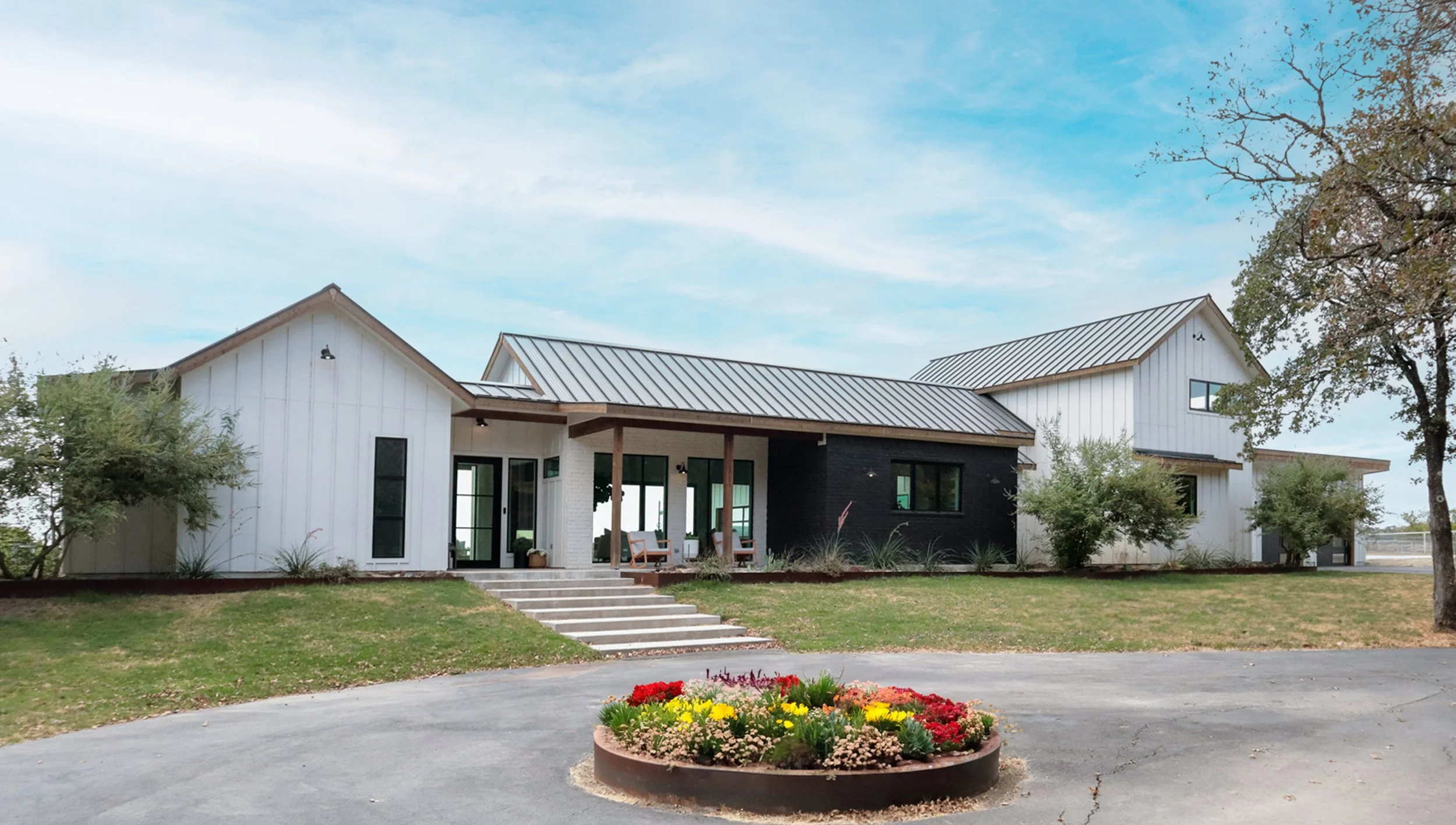A modern house with white and black exterior walls and a metal roof, surrounded by a well-maintained lawn and garden with colorful flowers in a circular flower bed, with trees and a partly cloudy sky in the background.