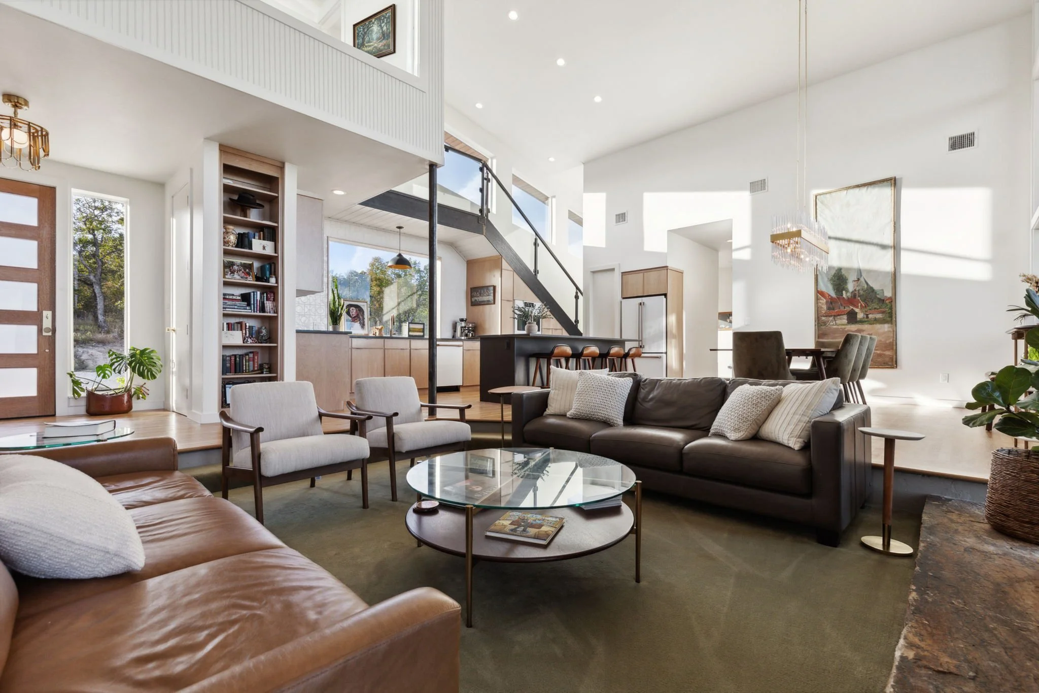 Open concept living room with large windows, white walls, wooden accents, a black staircase, and modern furniture including a brown leather sofa, white armchairs, and a round glass coffee table.