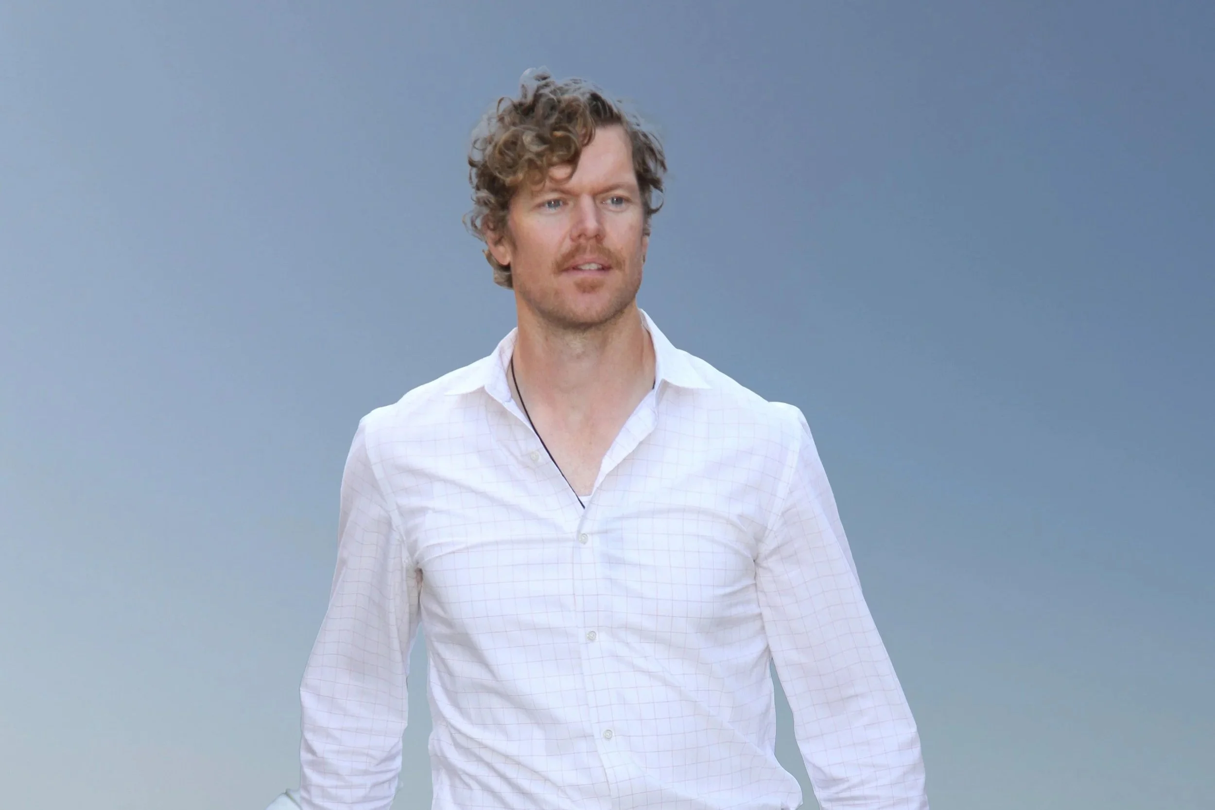 A man with curly hair, blue eyes, and a light beard wearing a white checkered shirt standing against a plain light blue background.