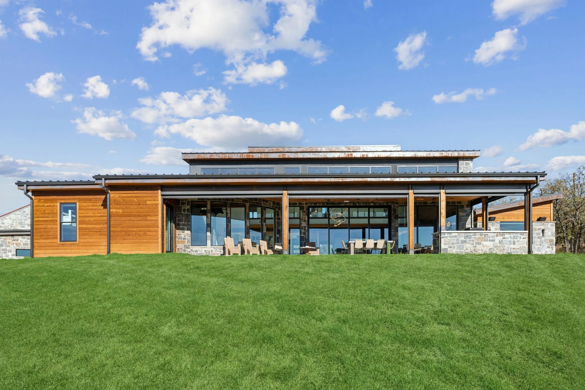 Modern house with large glass windows and a patio, set on a grassy lawn under a partly cloudy sky.