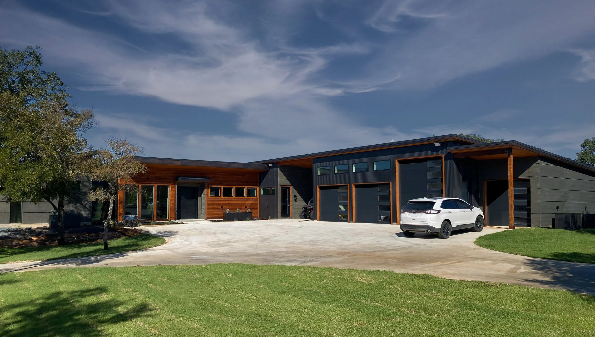 Modern house with a mix of wood and concrete exterior, parked white SUV in driveway, green lawn, trees, and a blue sky with clouds.