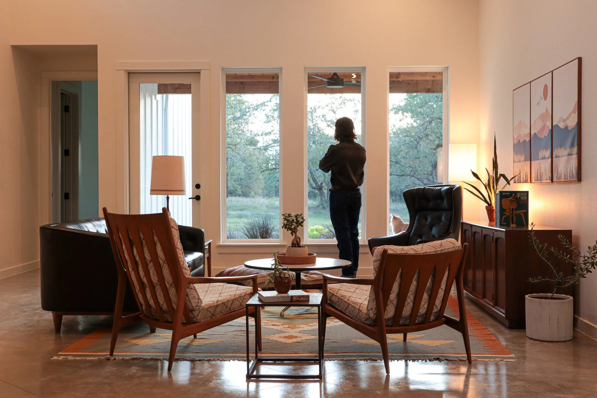 Living room with two armchairs and a sofa around a coffee table, a woman looking out the window, and a cat sitting on a chair, with trees visible outside.