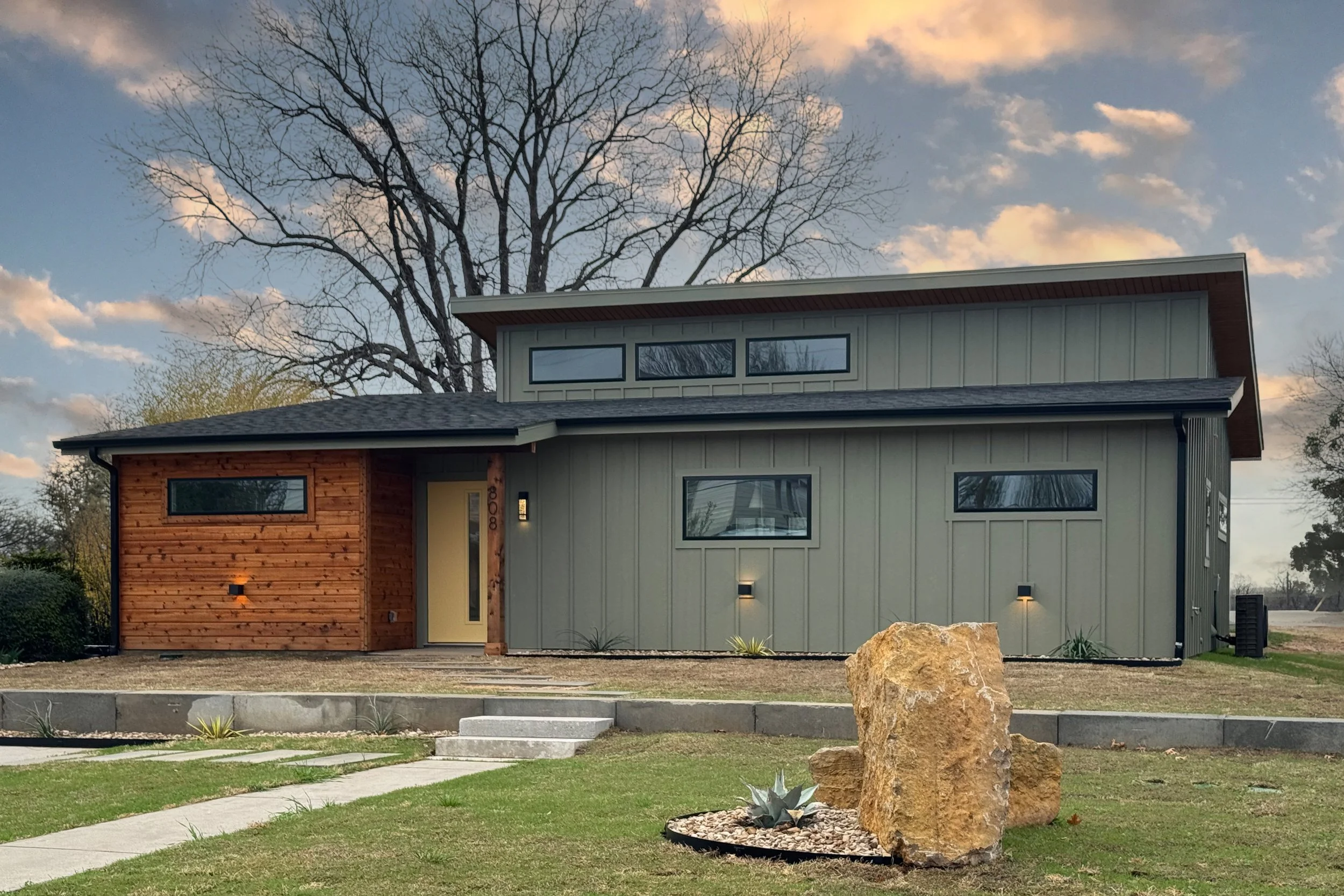 Modern two-story house with a mix of wood and gray siding, large windows, and a landscaped front yard with rocks and plants, under a cloudy sky.