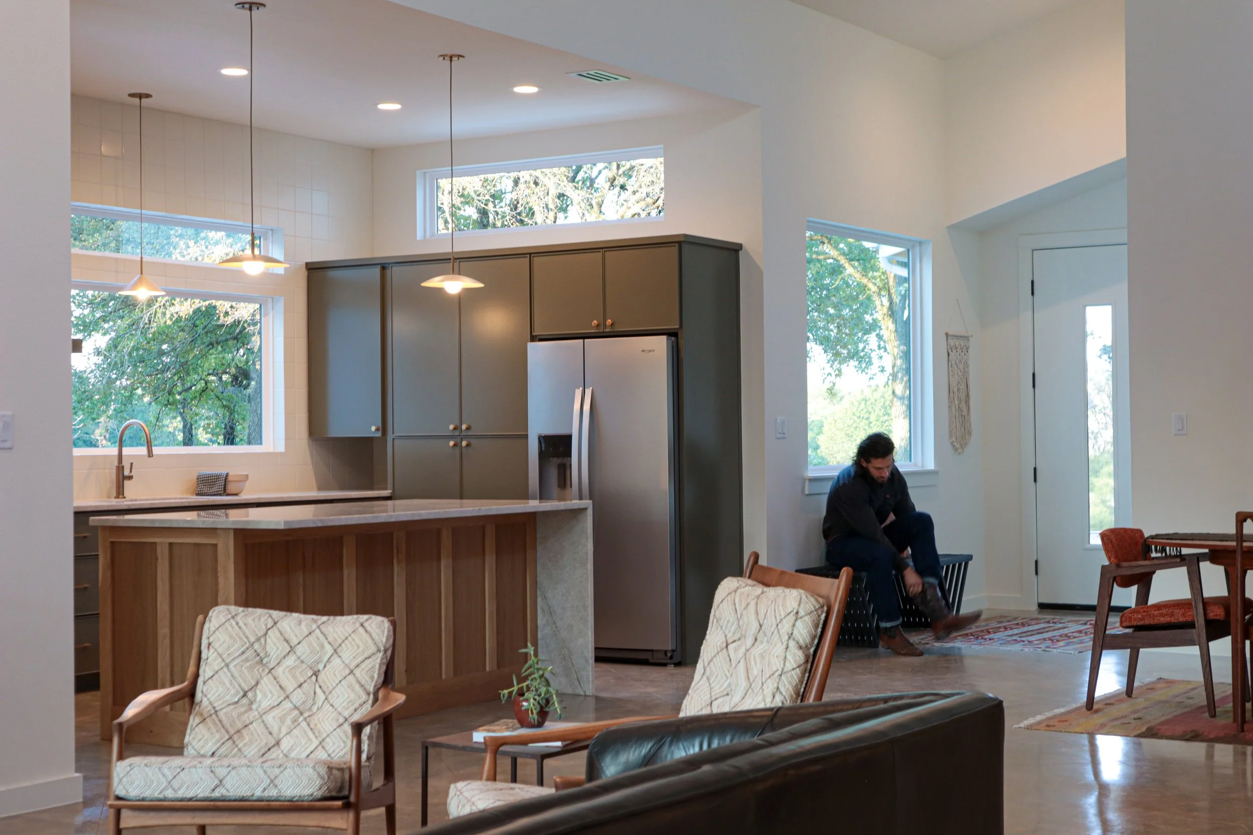 Interior of a modern open-concept living space featuring a kitchen with gray cabinets, a stainless steel fridge, a marble kitchen island, and large windows with views of trees. A person appears to be putting on shoes near the door.