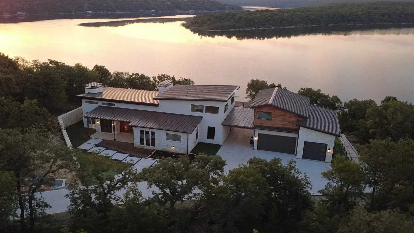 A modern house with white and wood exterior, metal roof, and large windows, situated on a hillside near a body of water during sunset, surrounded by trees.