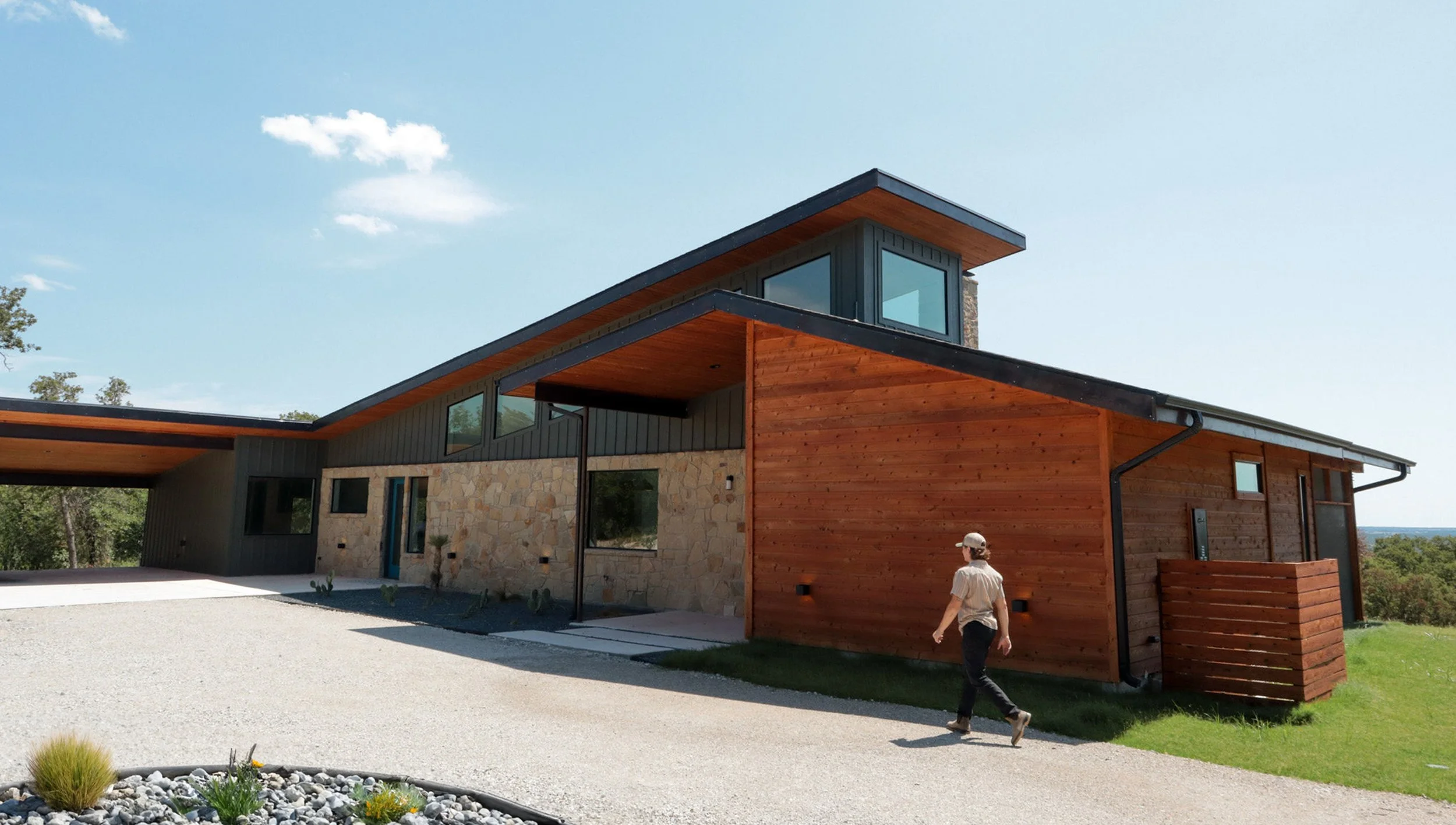 Modern house with stone and wood exterior, large windows, and sloped roof, on a clear day with a person walking nearby.