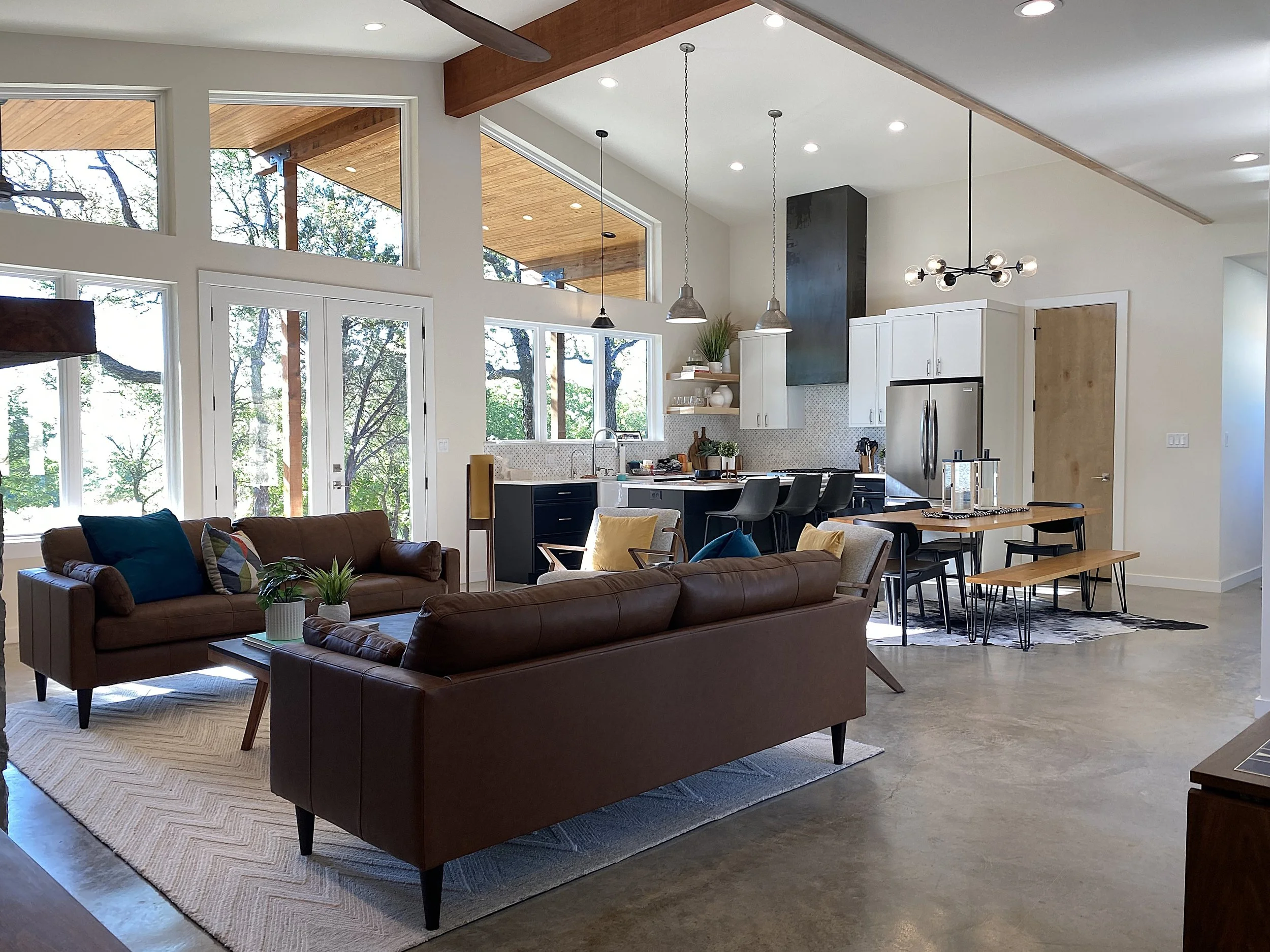 Open-concept living and kitchen area with large windows, brown leather sofas, a wooden dining table with black chairs, modern kitchen with white cabinets, stainless steel appliances, and a view of trees outside.