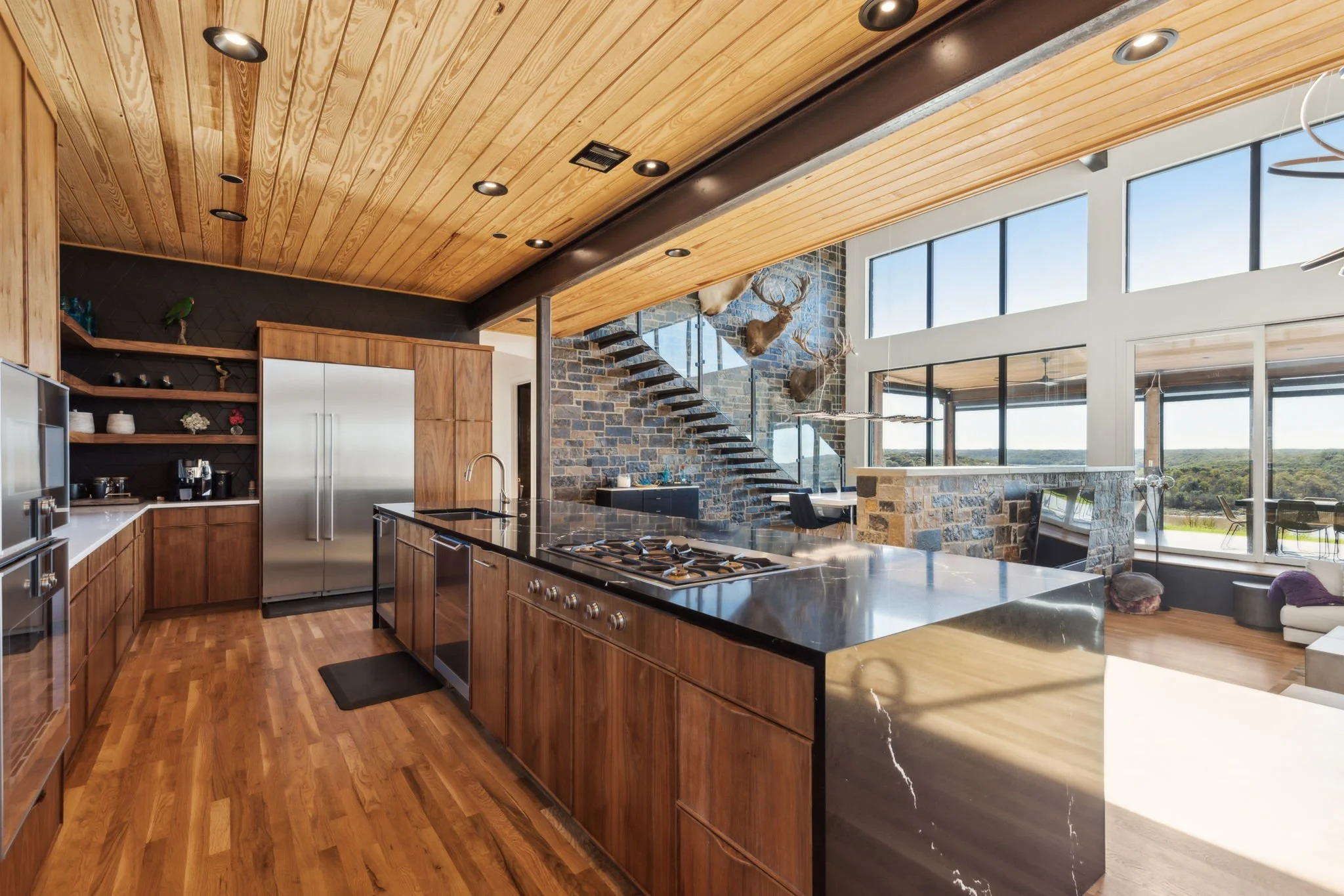 Open-concept modern kitchen with wooden cabinetry, black countertop, stainless steel appliances, and large floor-to-ceiling windows overlooking a scenic view, with a stone accent wall and mounted deer heads.