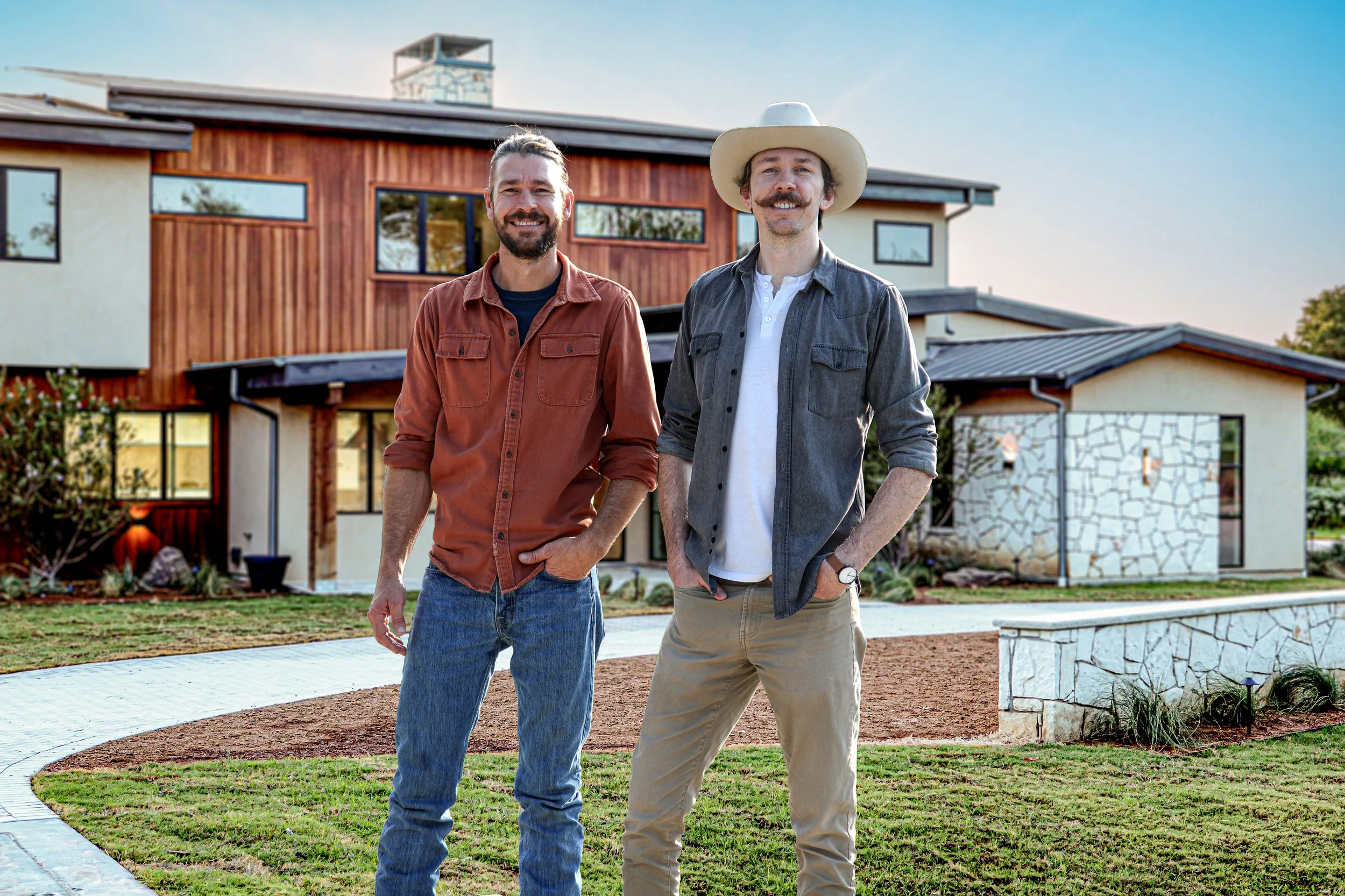Two men standing outdoors in front of a modern house with large windows and a wooden and stone exterior. One man wears a rust-colored shirt and jeans, smiling with his hands near his pockets. The other man wears a cowboy hat, gray jacket, and beige pants, also smiling. The background features a lawn, garden bed, and a clear blue sky.