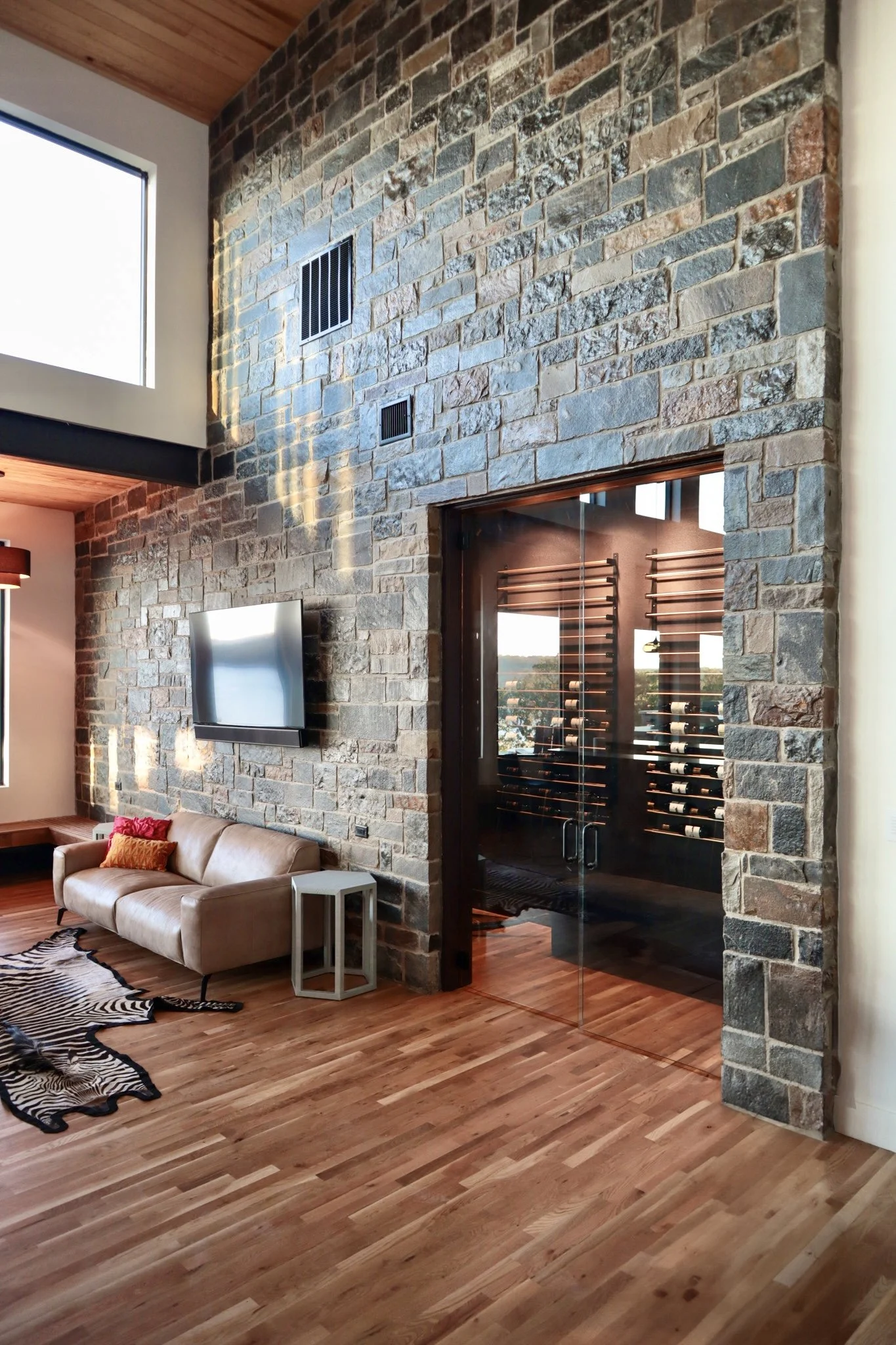 Living room with a stone accent wall, flat screen TV, beige sofa with pillows, a zebra print rug, wooden flooring, and a glass door leading to a wine cellar filled with wine bottles.