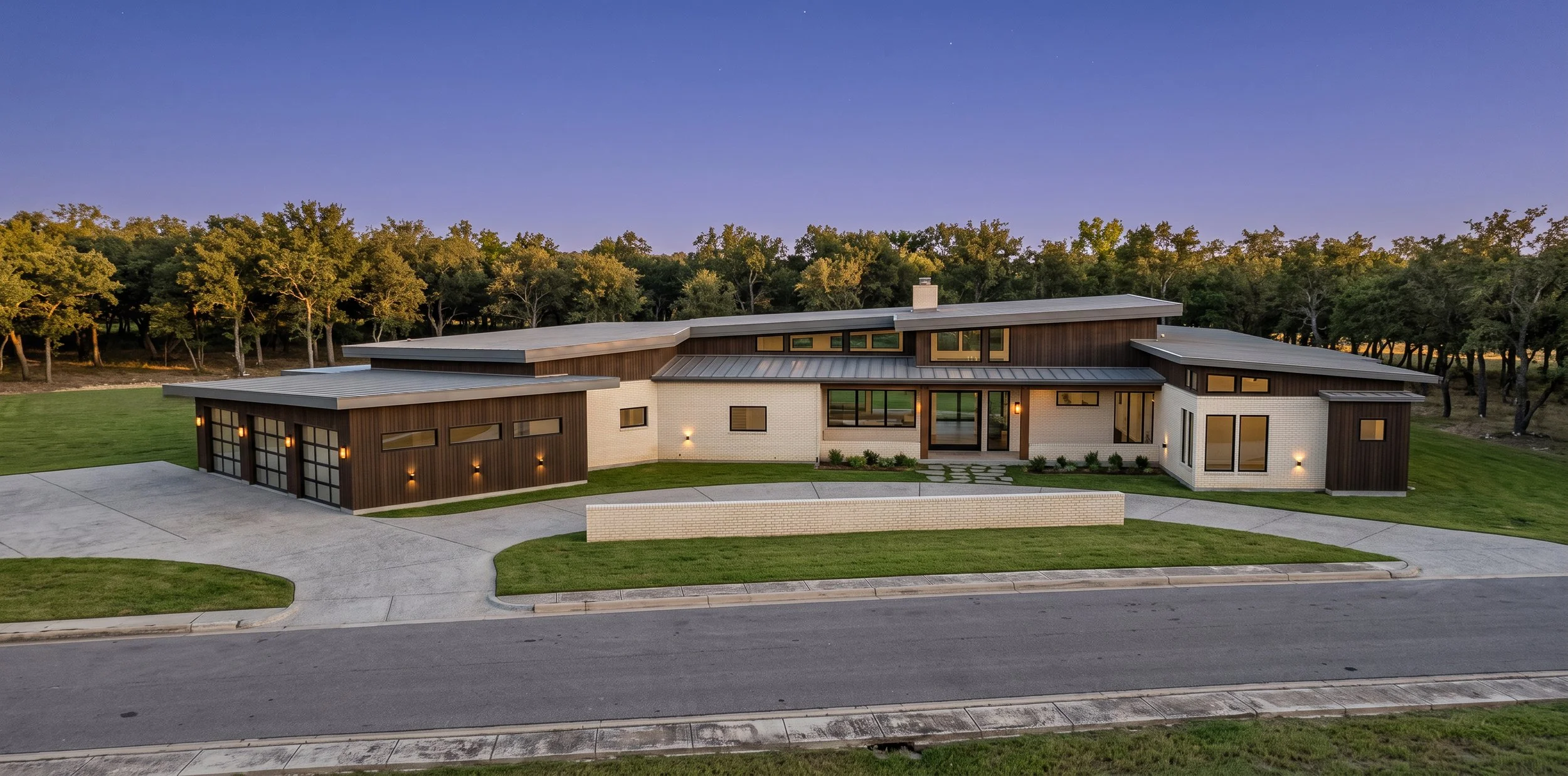 Modern house with a mix of white brick and dark wood exterior, large windows, and a multi-level metal roof, surrounded by a well-maintained lawn and trees at sunset.