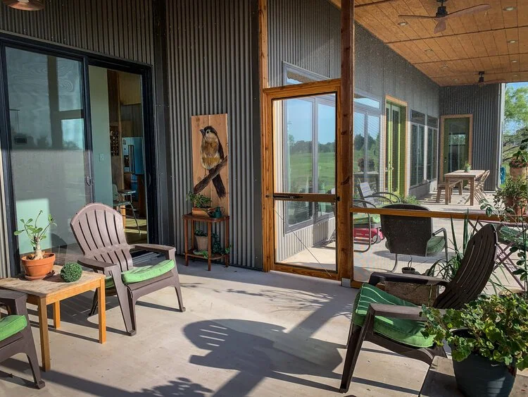 A covered porch with outdoor chairs, potted plants, and a bird mounted on the wall, overlooking a grassy field.