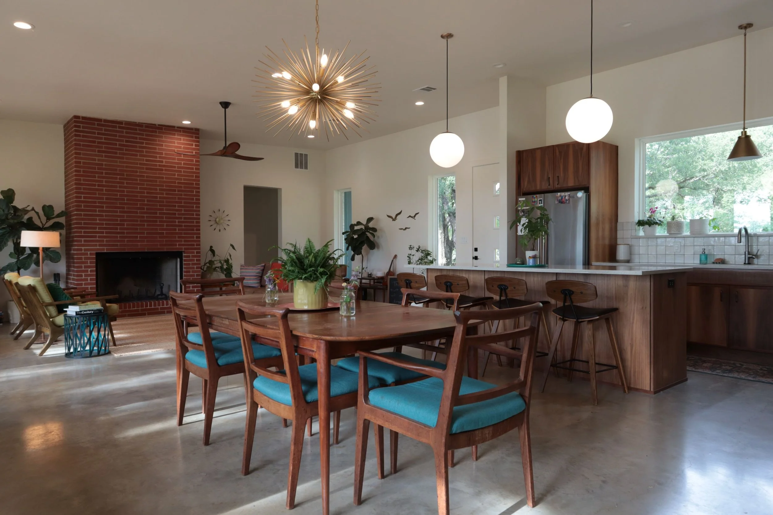 Open concept living and kitchen area with a fireplace, dining table with teal cushions, plants, pendant and chandelier lights, and large windows with greenery outside.