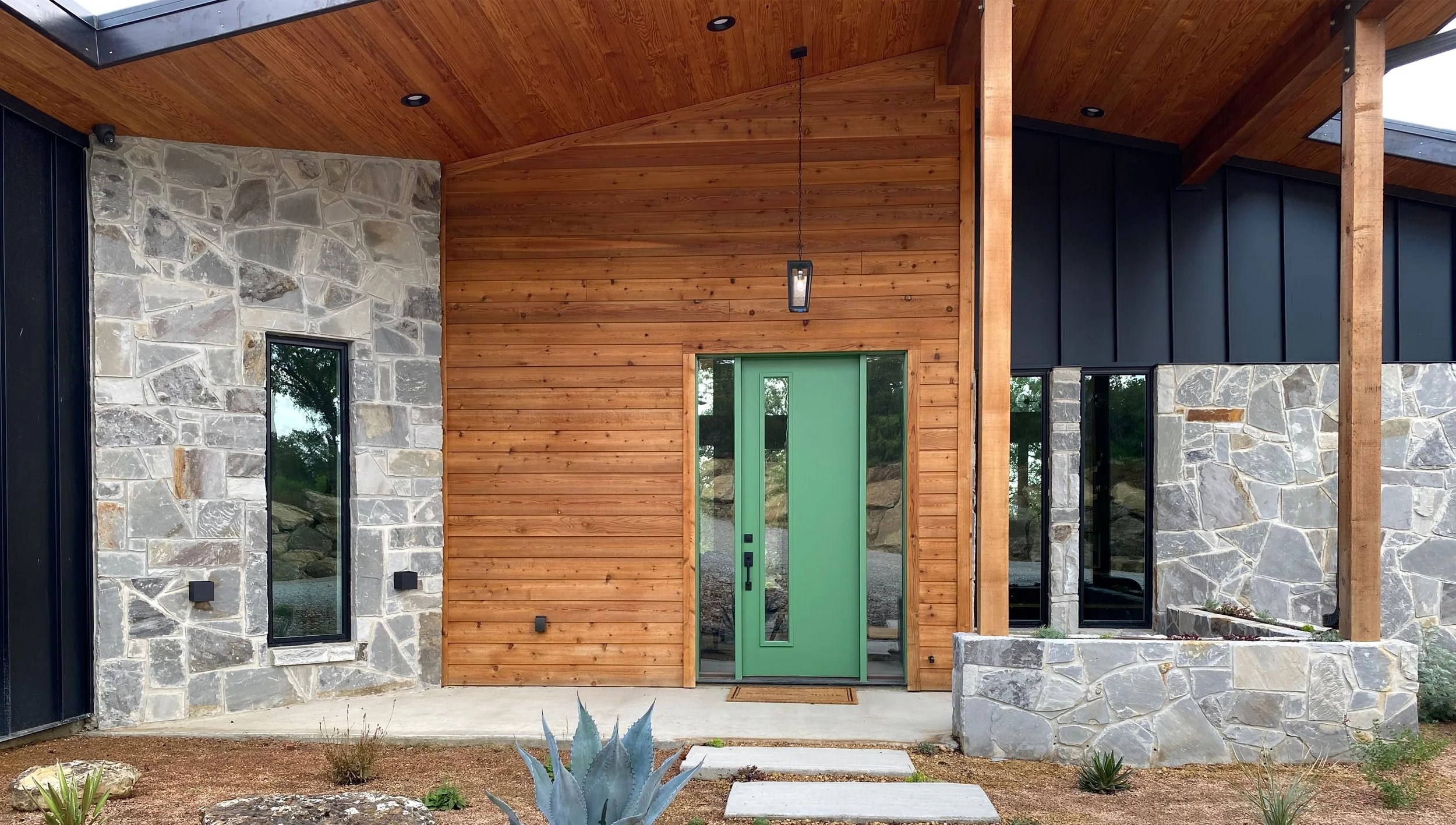 Modern house exterior featuring a green door, stone and wood siding, with a pathway and desert plants in the front yard.