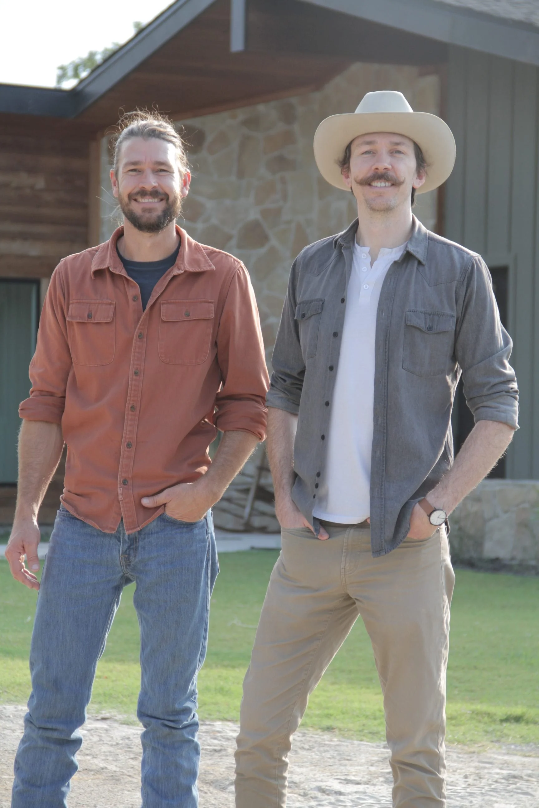 Two smiling men standing outdoors in front of a modern house, dressed casually. One man is wearing a rust-colored shirt and jeans, and the other is wearing a gray shirt, khaki pants, and a cowboy hat.