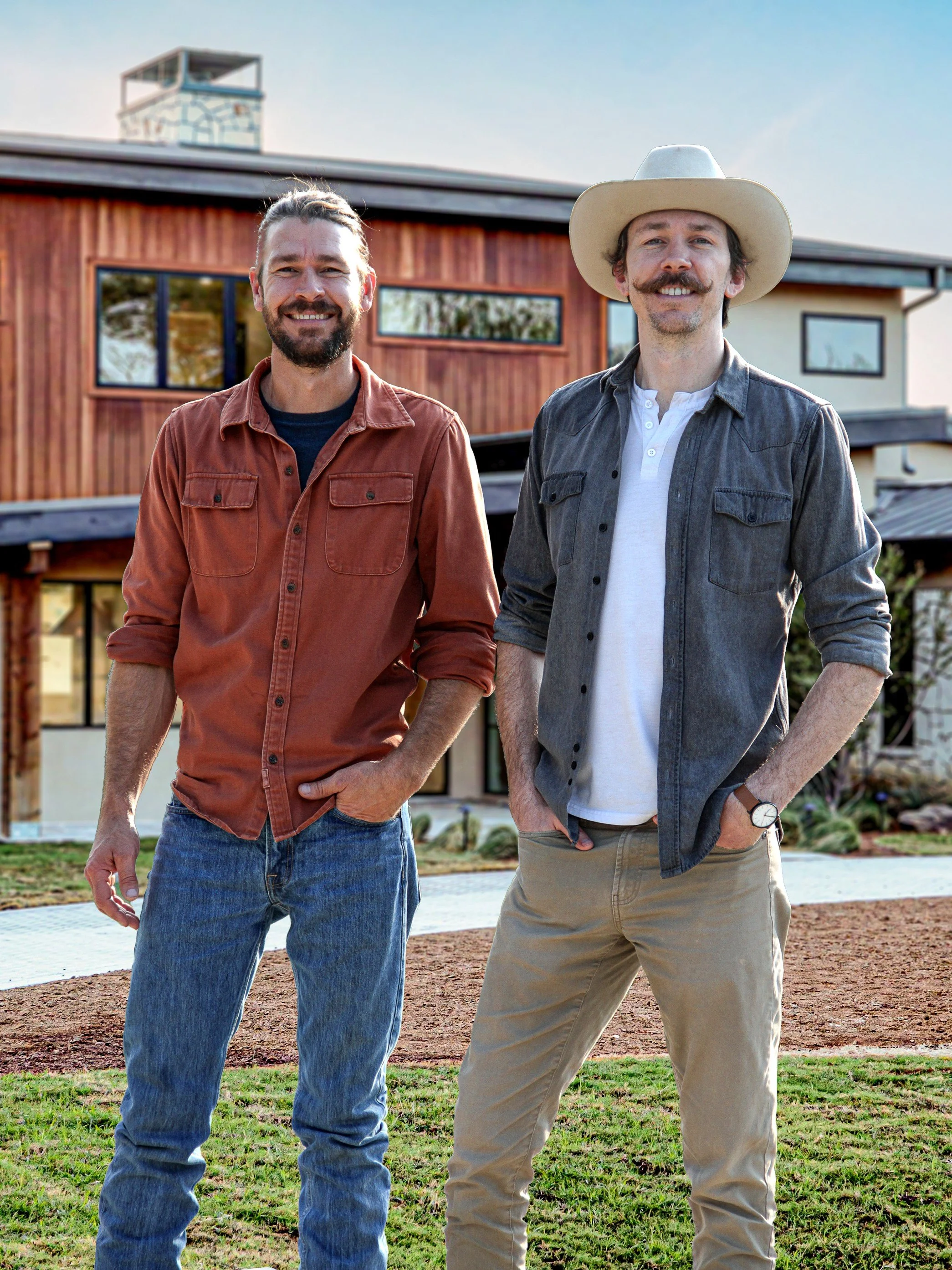 Two young men standing on a lawn in front of a modern house, both smiling. One has a beard and is wearing a brown shirt and jeans; the other has a mustache and is wearing a wide-brimmed hat, a gray shirt, and khaki pants.