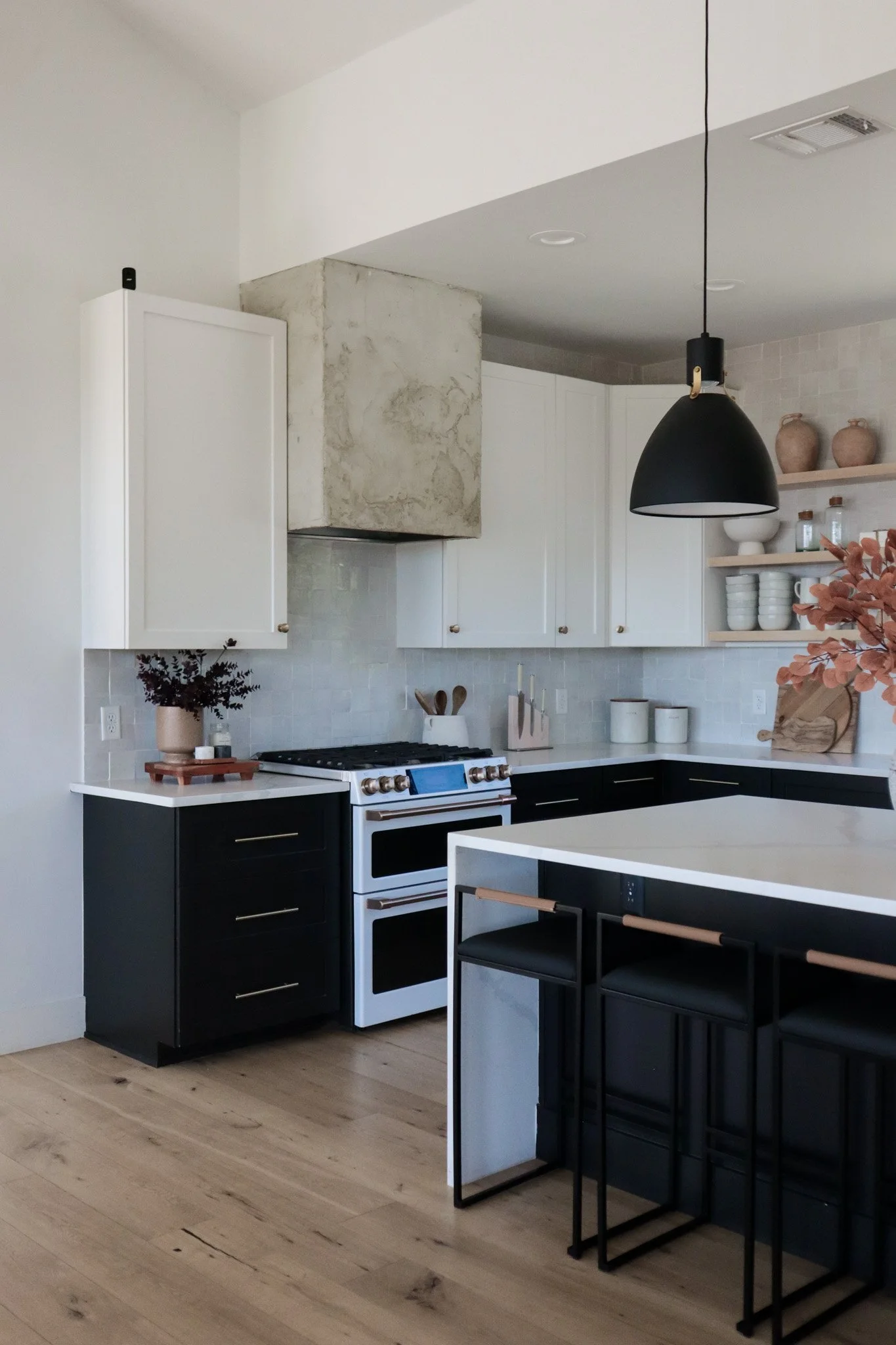Modern kitchen with white upper cabinets, black lower cabinets, a white countertop, a black pendant light, a stove, open shelves with ceramics, and bar stools at the island.