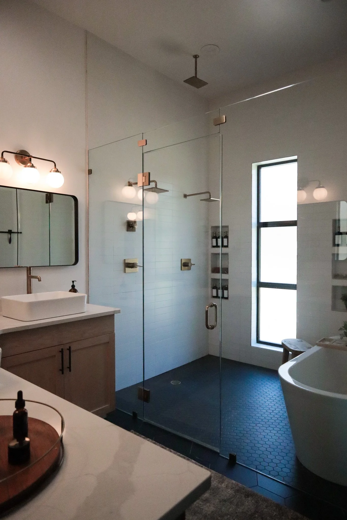 Modern bathroom with glass shower enclosure, white sink vanity, and a large window, with black hexagonal floor tiles and minimal decor.